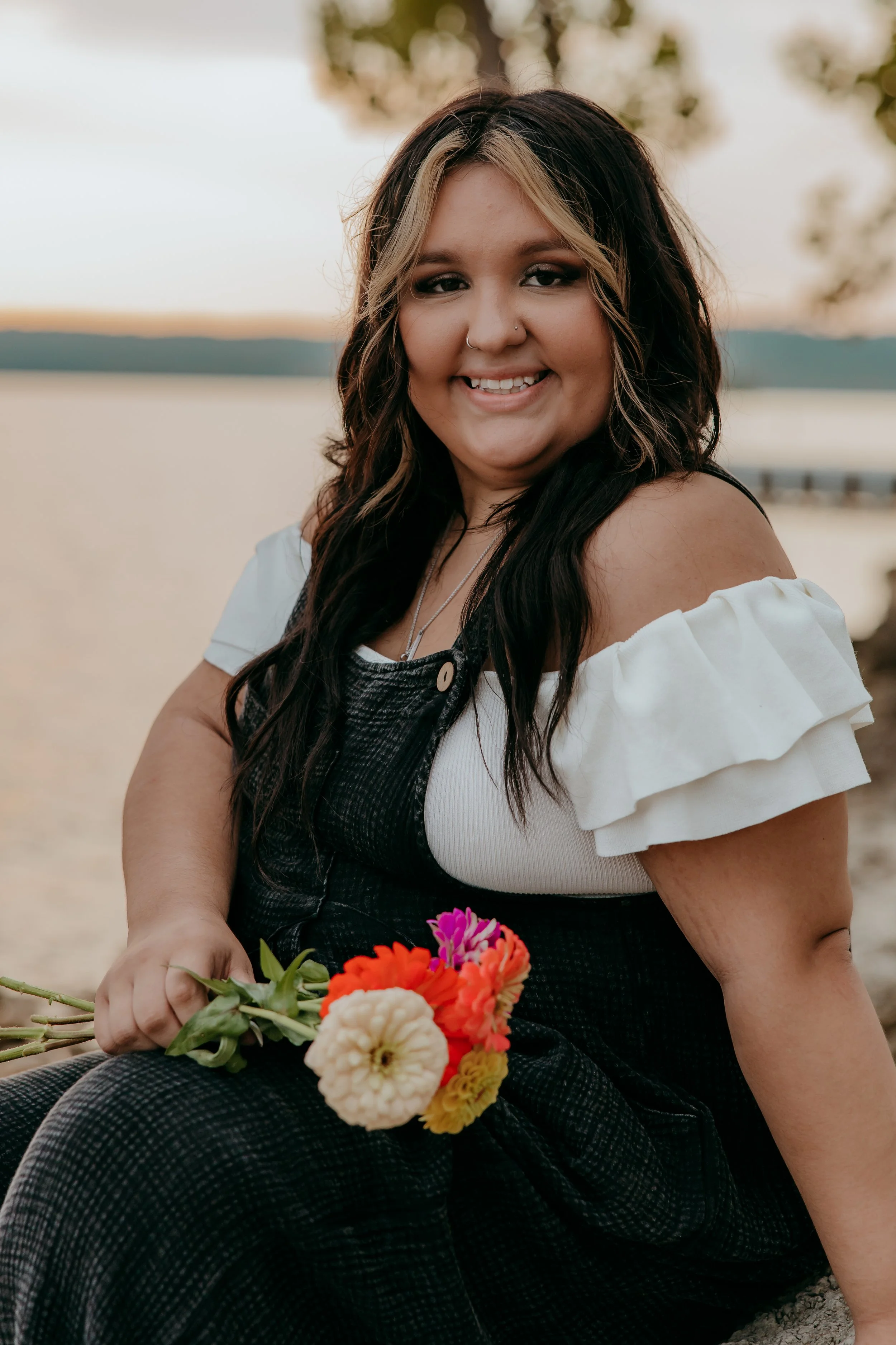 A woman sitting on the ground near a body of water holding a small bouquet of colorful flowers, with trees and water in the background at sunset.
