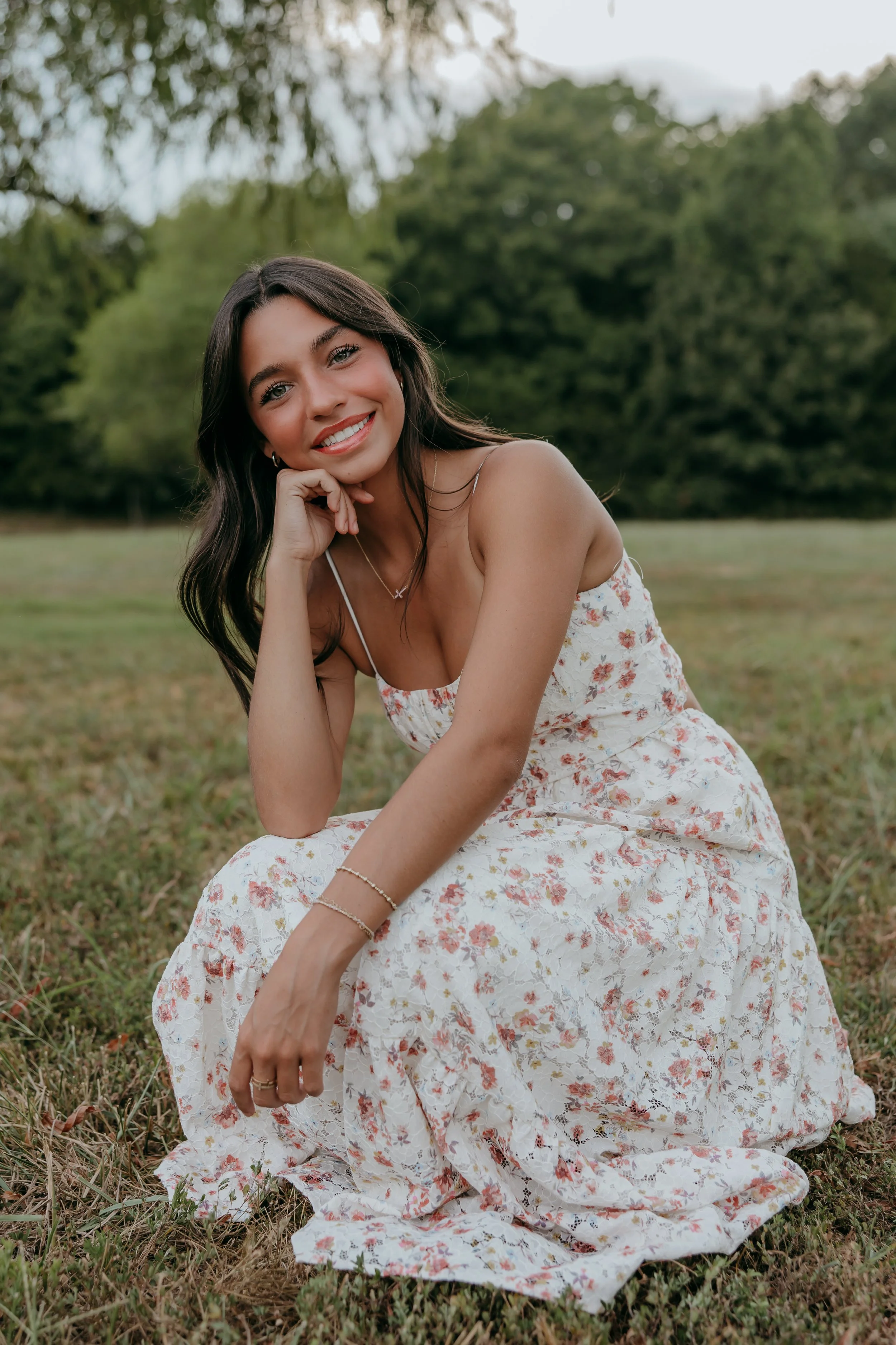 A young woman with long dark hair, wearing a white floral dress, is sitting on the grass in an outdoor park setting, smiling at the camera with a relaxed pose and her chin resting on her hand.