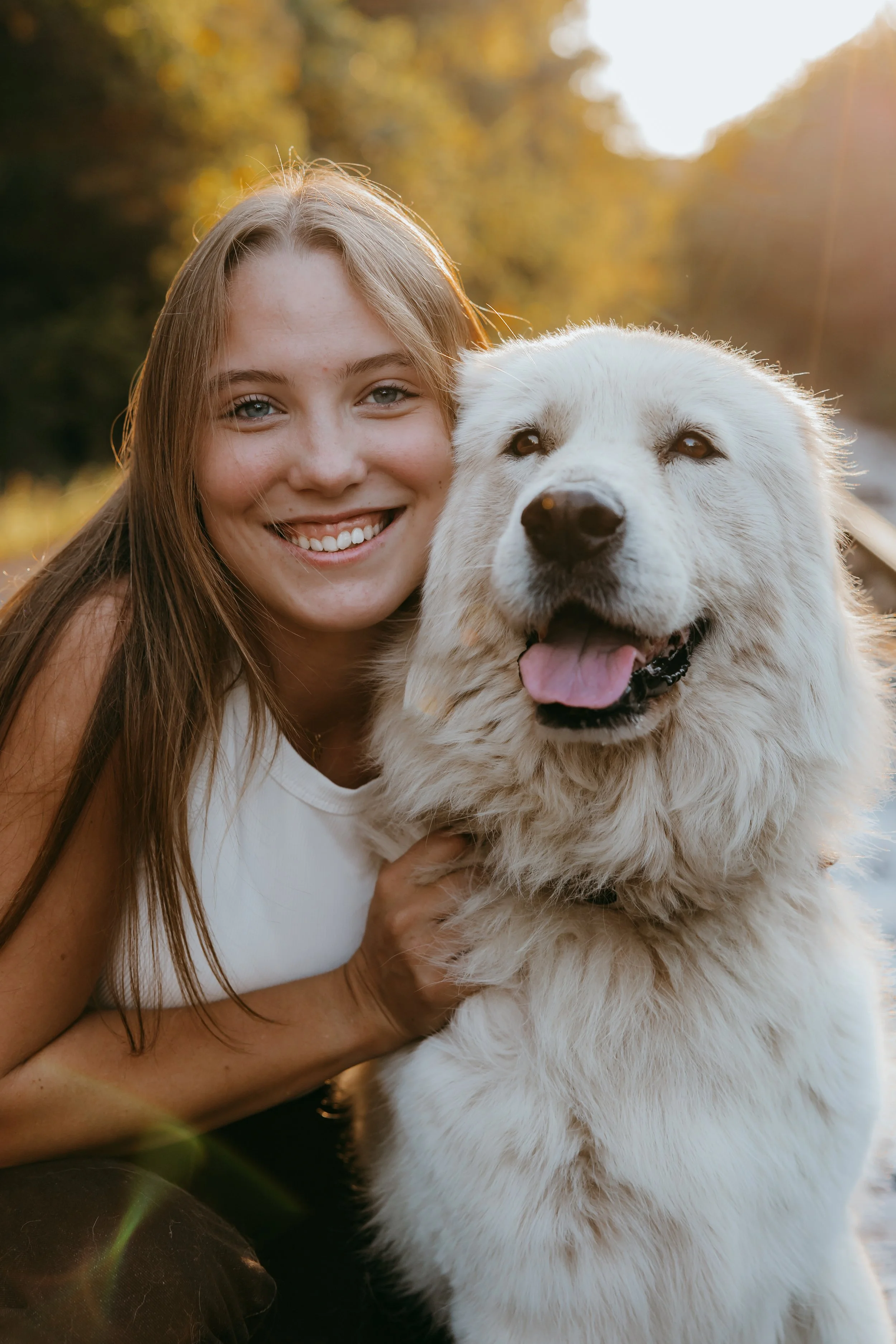 A young woman smiling and holding a large white dog outdoors during sunset with trees in the background.
