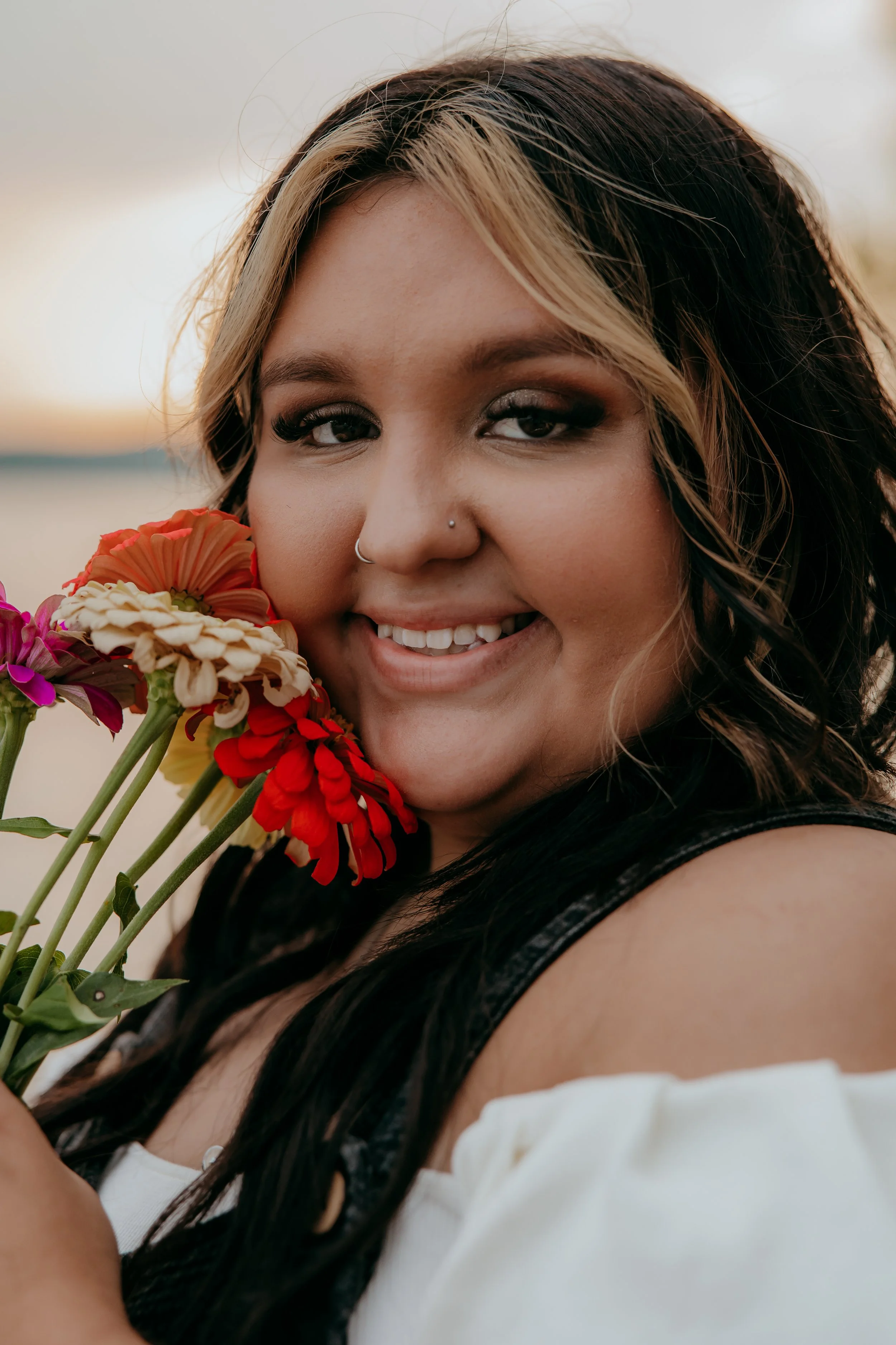 A woman with long dark hair and blonde highlights holding a bouquet of colorful flowers near her face, smiling at the camera outdoors during sunset.