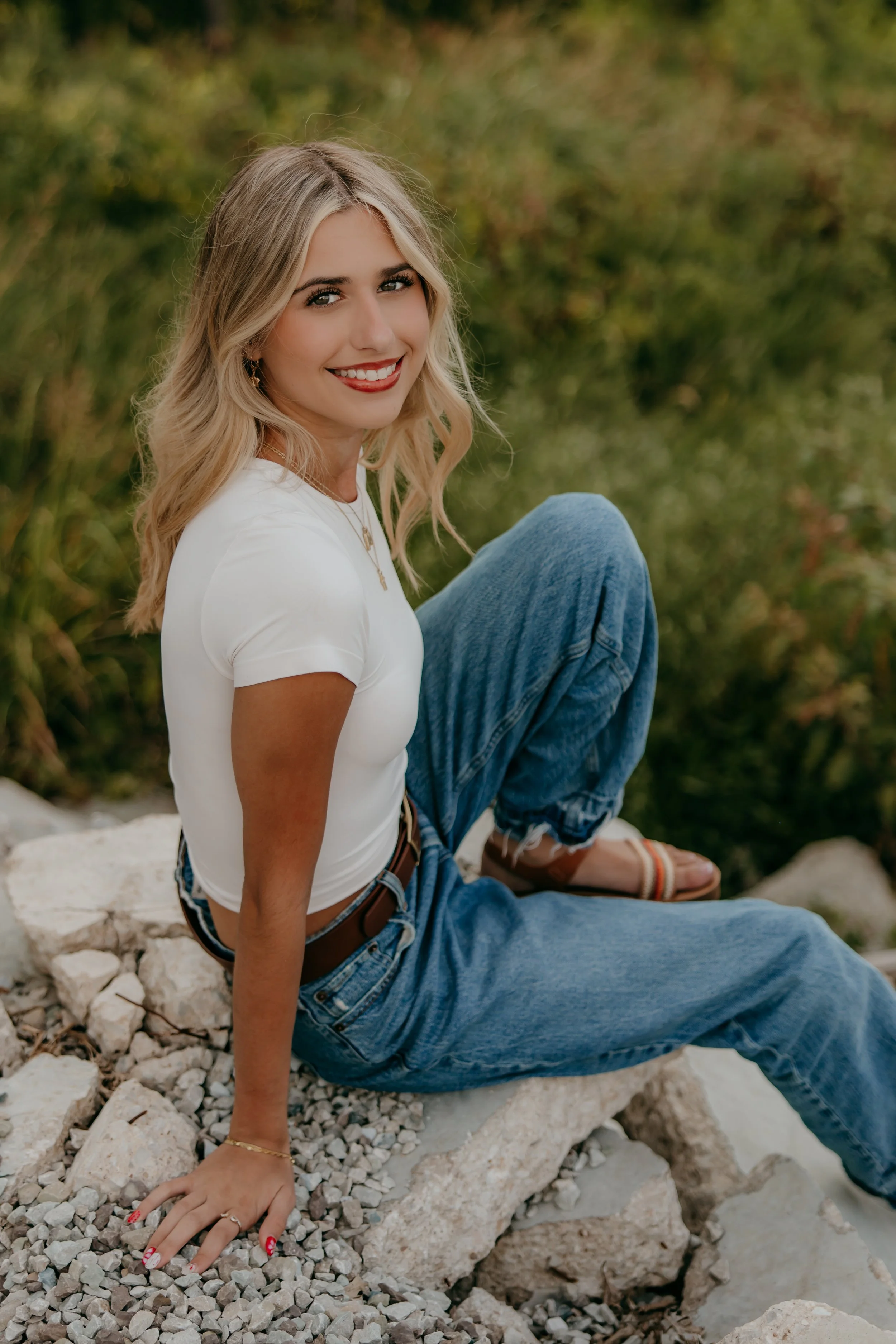 A smiling young woman with blonde, wavy hair sitting on rocks outdoors in a natural setting with greenery.