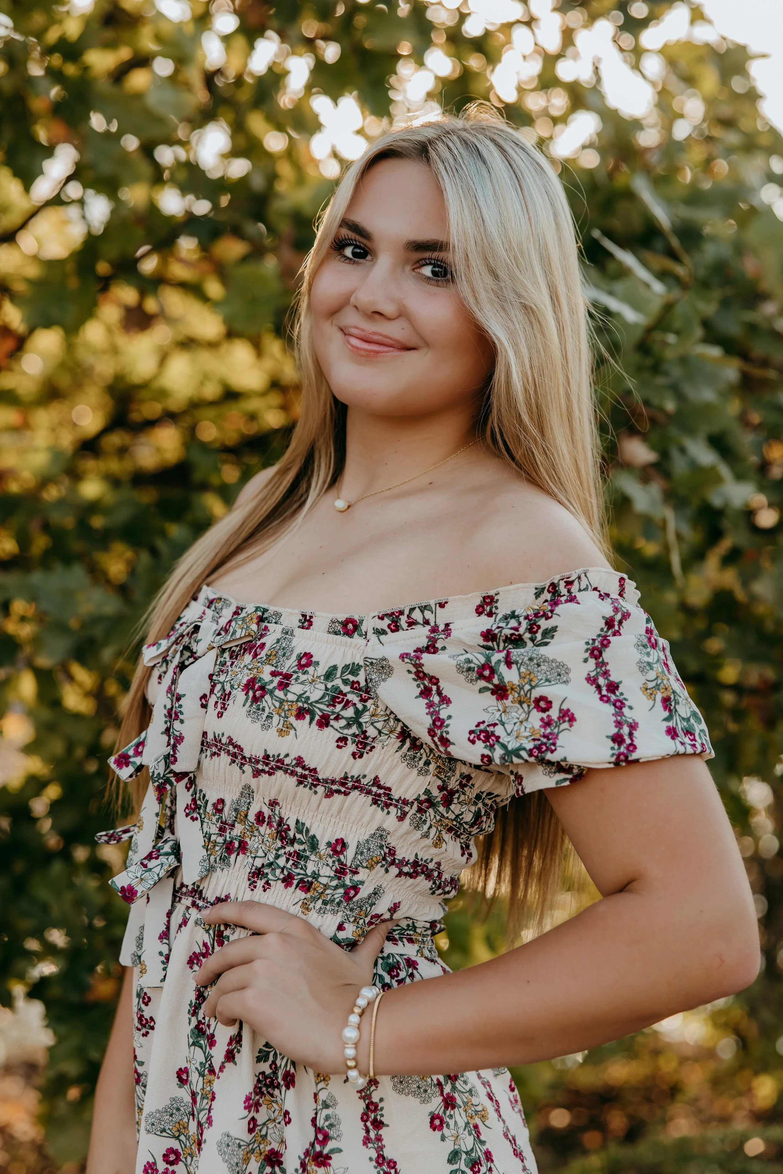 A young woman with long blonde hair, wearing a floral off-the-shoulder dress, standing outdoors with green leafy background, smiling at the camera.