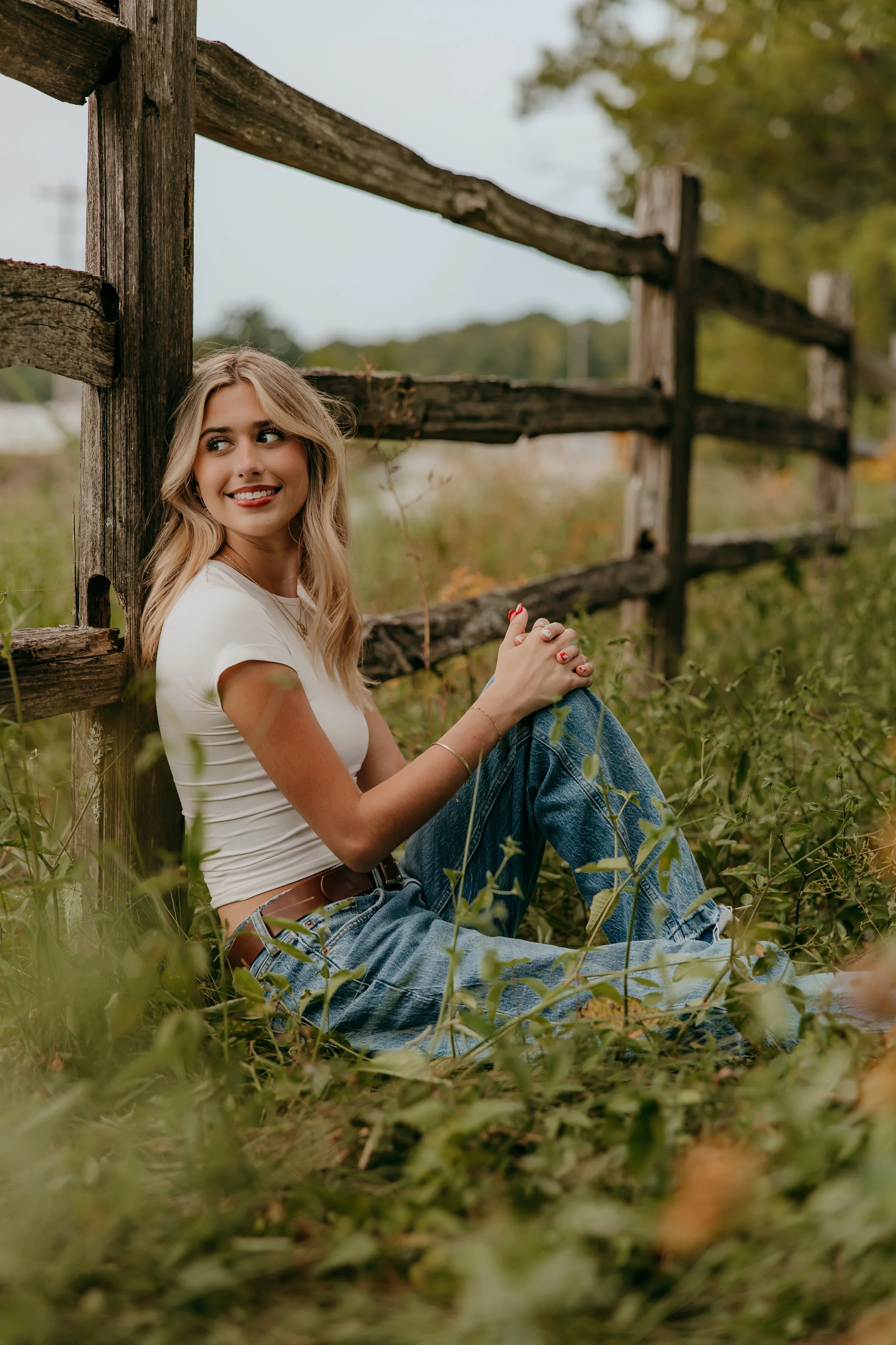 A young woman with blonde hair, sitting on the ground near a wooden fence in a grassy field, smiling and looking to the side.