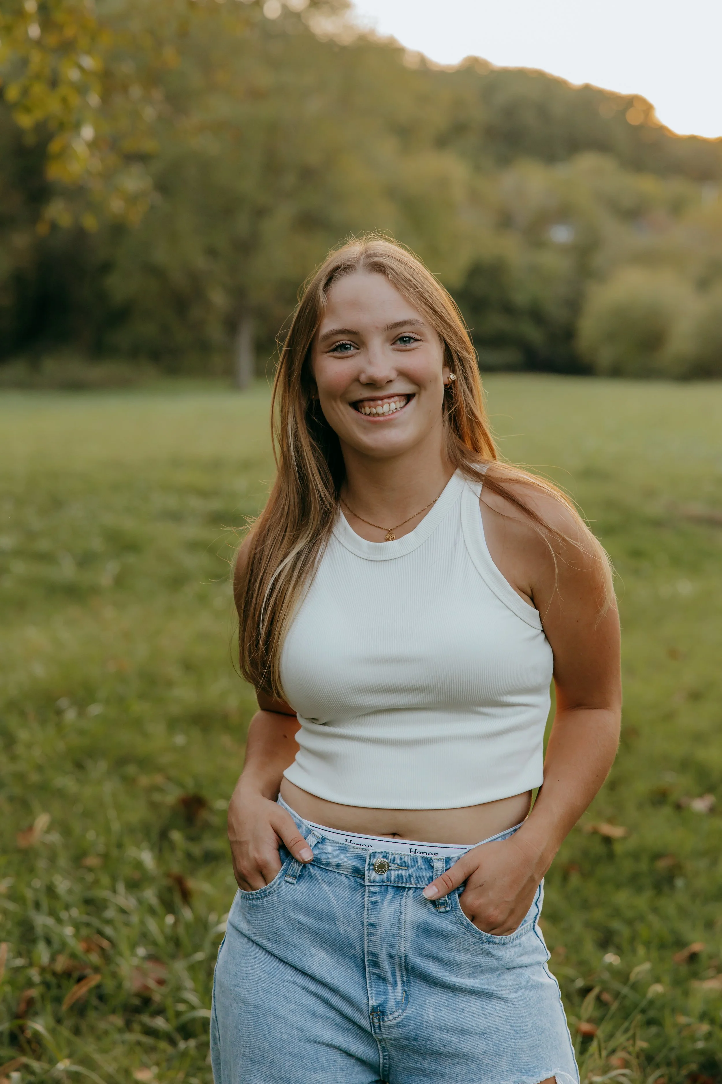 A young woman with long, light brown hair standing outdoors in a grassy area with trees in the background, smiling at the camera, wearing a white sleeveless top and light blue high waisted jeans.