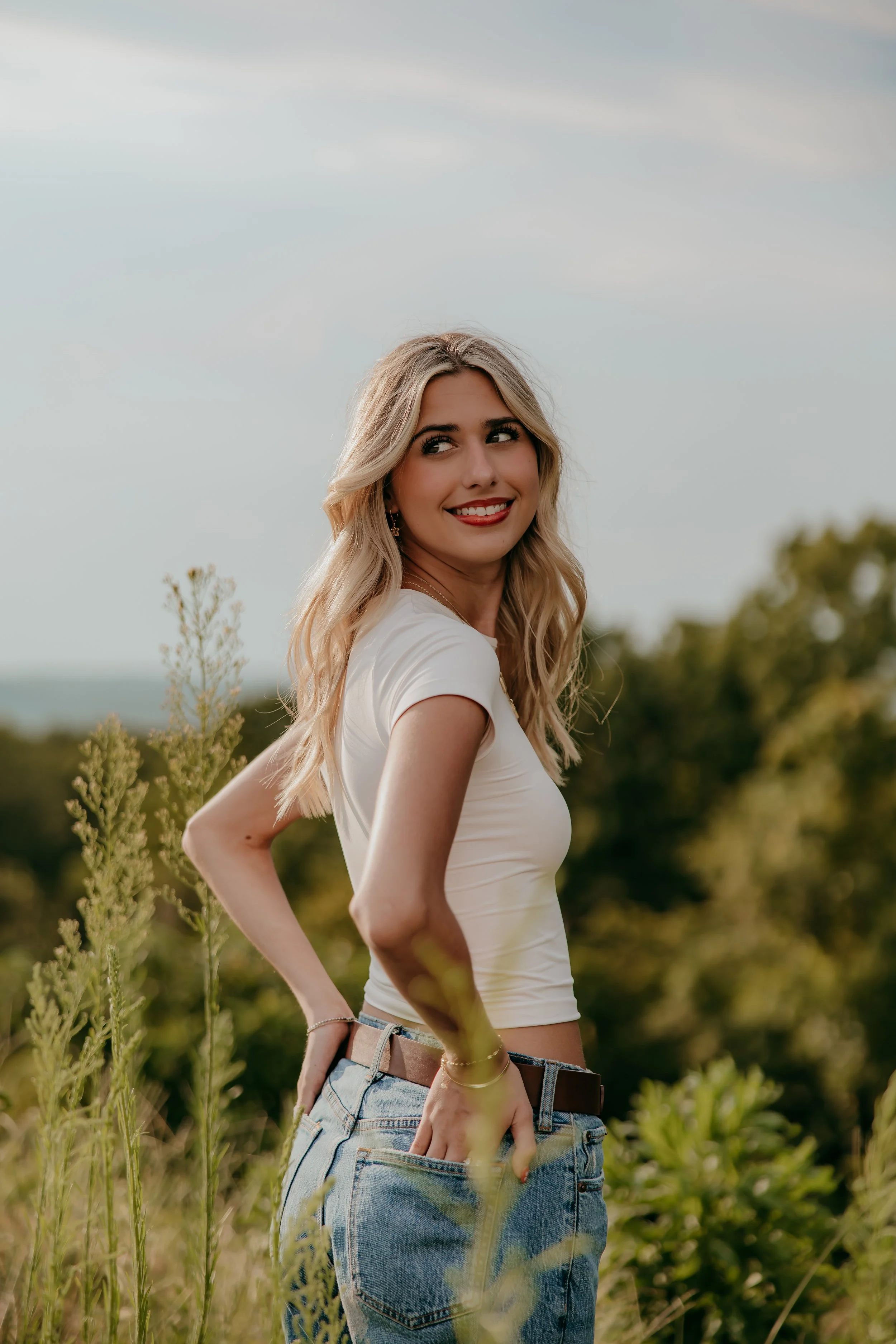 A young woman with blonde hair, wearing a white t-shirt and blue jeans, standing outdoors among tall grass and trees, smiling and looking to her left.