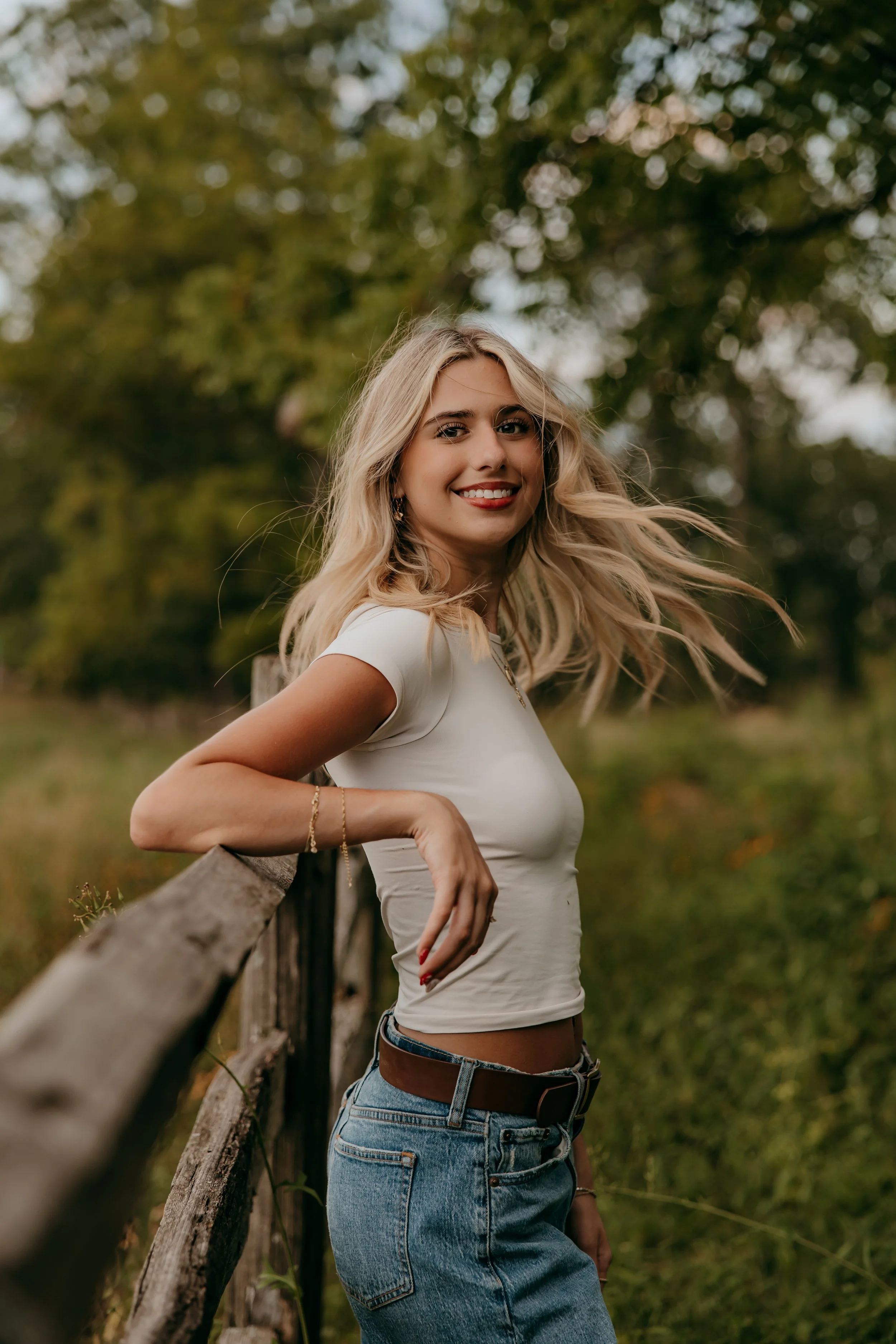 A young woman with long blonde hair, wearing a white crop top and blue jeans, leaning on a wooden fence outdoors surrounded by greenery, smiling at the camera.