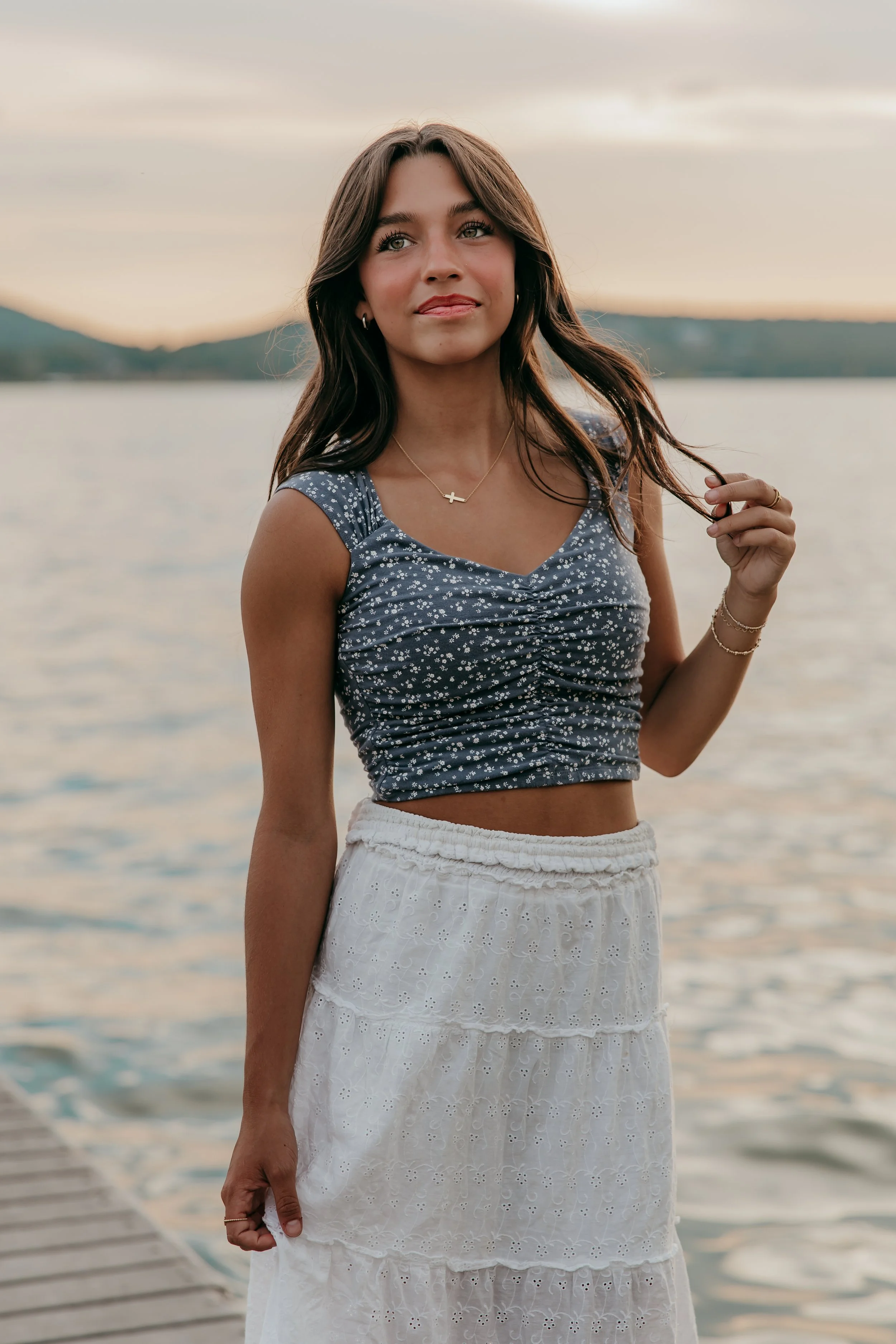 A young woman stands on a dock by a body of water at sunset, holding a lock of her long brown hair with her right hand, wearing a navy blue sleeveless crop top with white floral pattern and a white tiered skirt.