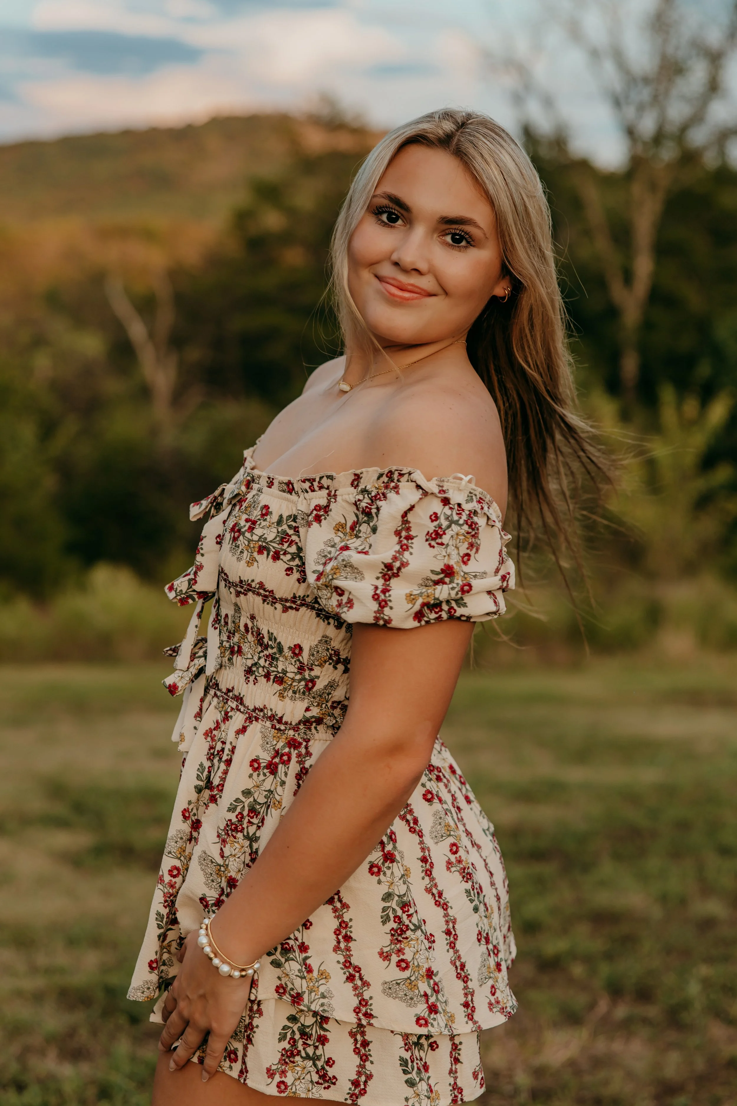 A young woman with light brown hair, smiling, standing outdoors in a field with trees and a hill in the background, wearing a floral off-the-shoulder dress.