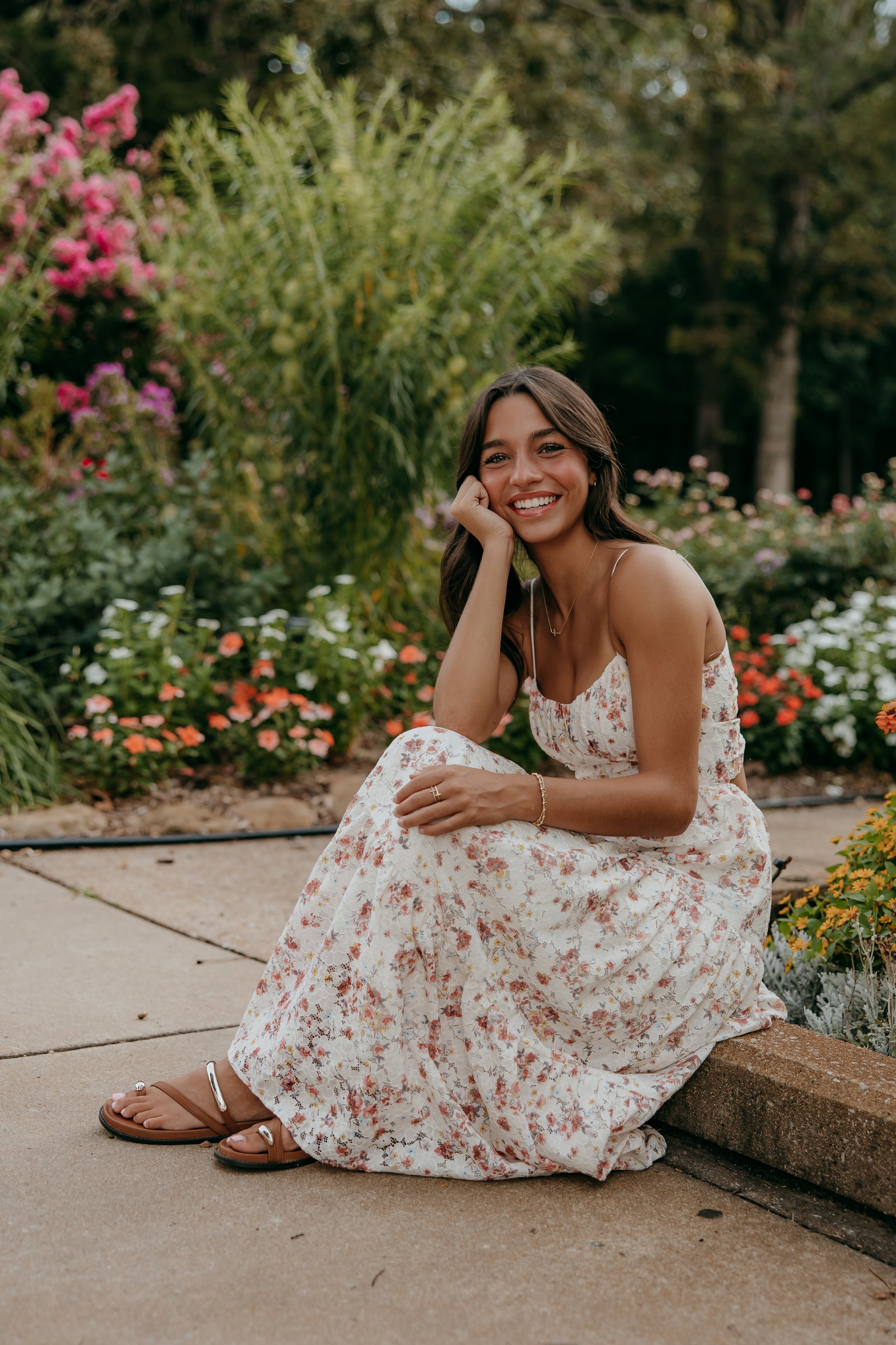 A young woman with long dark hair, wearing a sleeveless floral dress and brown sandals, sitting on a low brick wall in a garden with colorful flowers and green foliage, smiling at the camera.