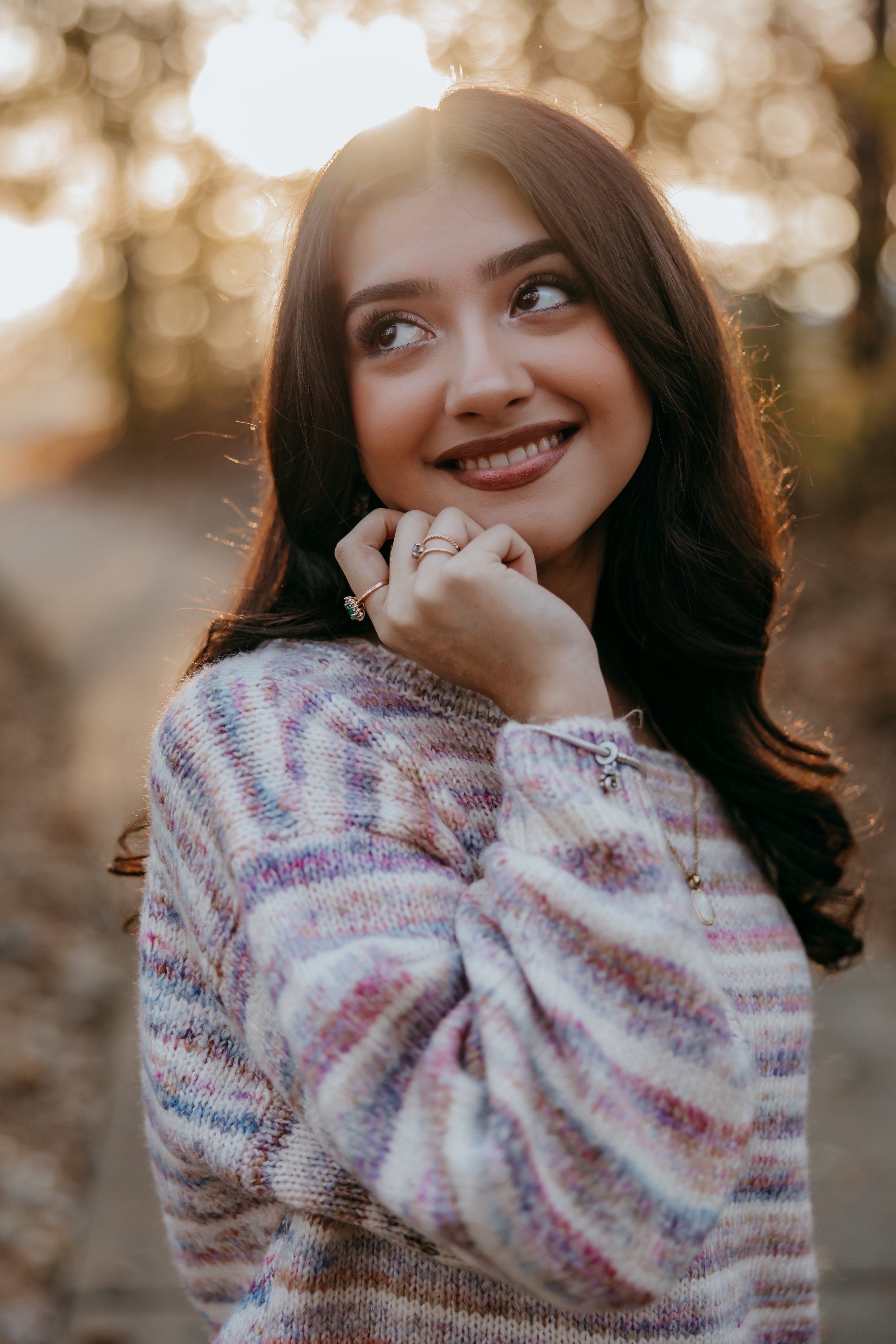 Young woman outdoors in autumn, smiling, wearing a colorful sweater, with sunlight behind her.