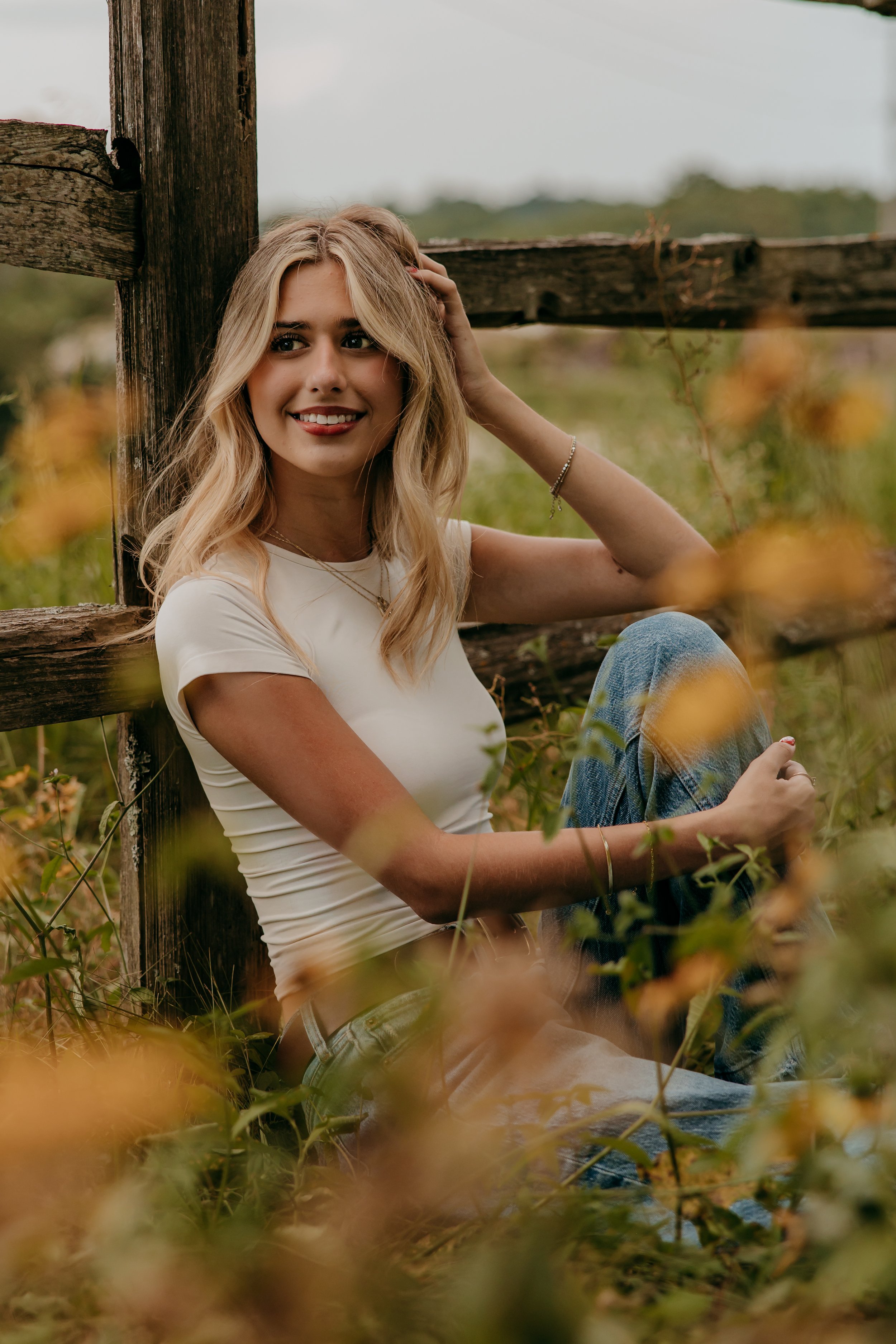 A young woman with blonde hair sitting outdoors near a wooden fence, smiling, with a field of yellow flowers.