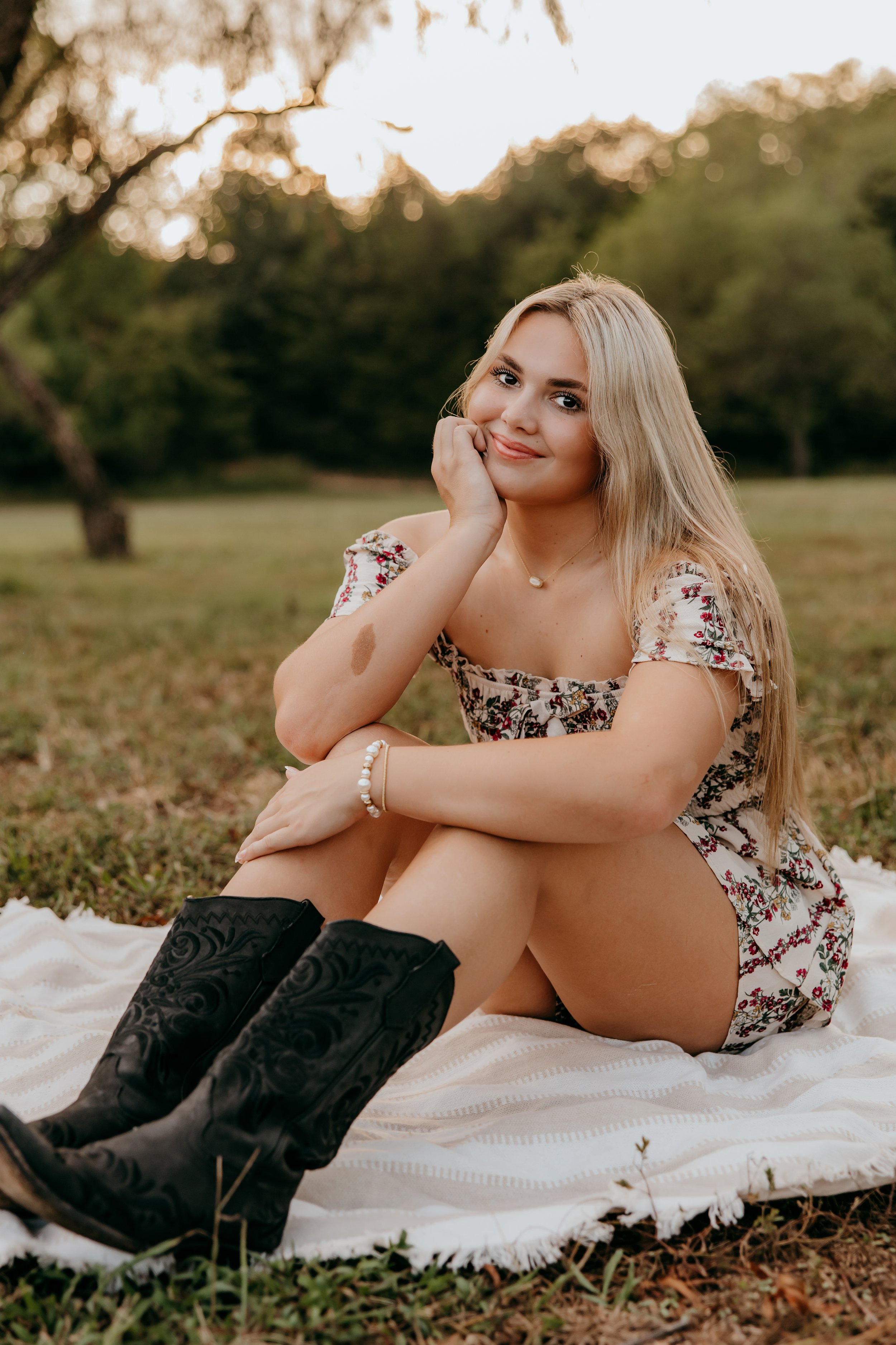 A young woman with long blonde hair sitting on a white blanket outdoors in a park during the late afternoon or early evening. She is resting her face on her hand, smiling, and wearing a floral dress, black cowboy boots, and jewelry. The background fe