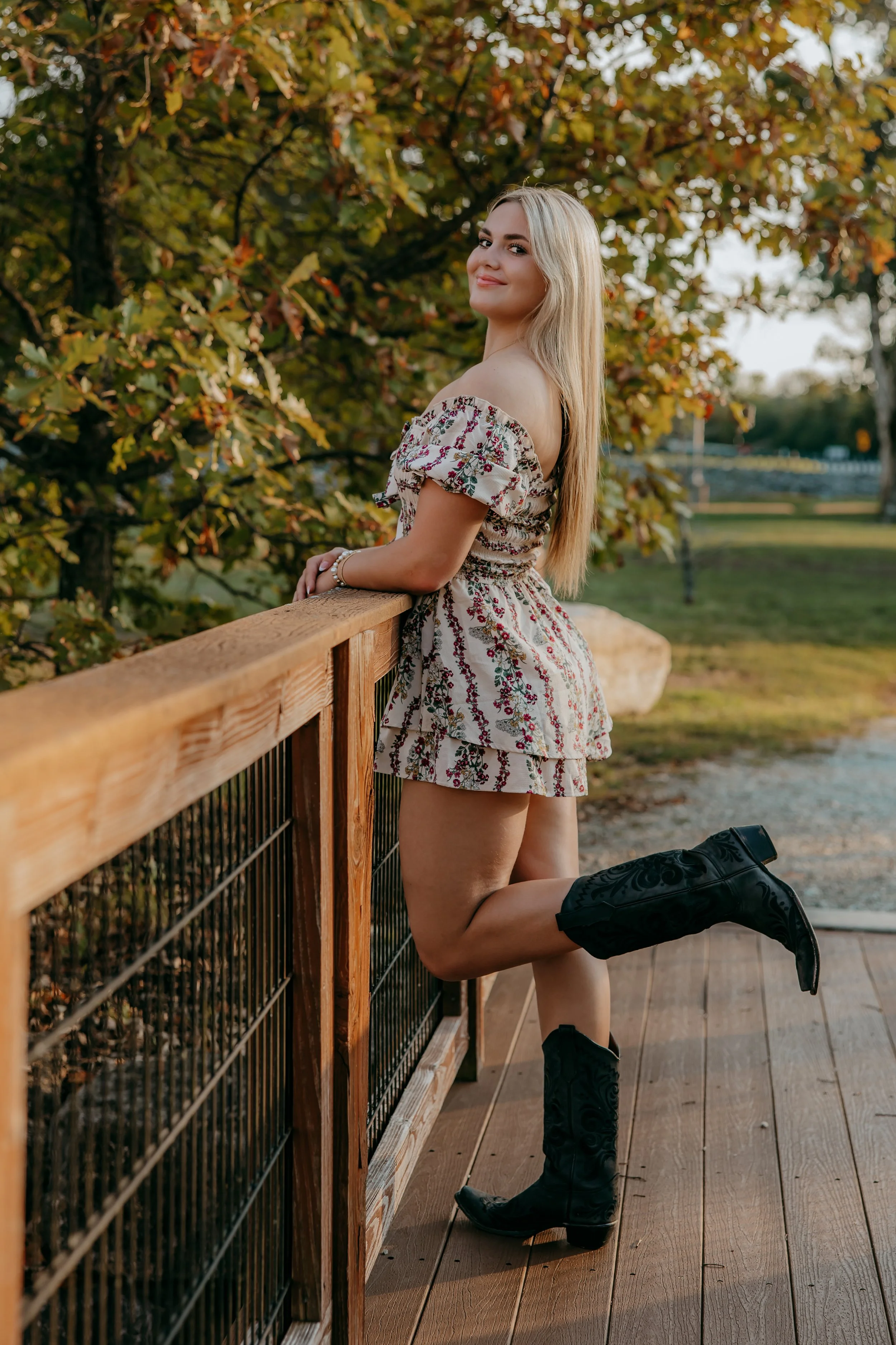 A young woman with long blonde hair wearing a patterned dress and black cowboy boots, leaning on a wooden railing outdoors in a park during fall.