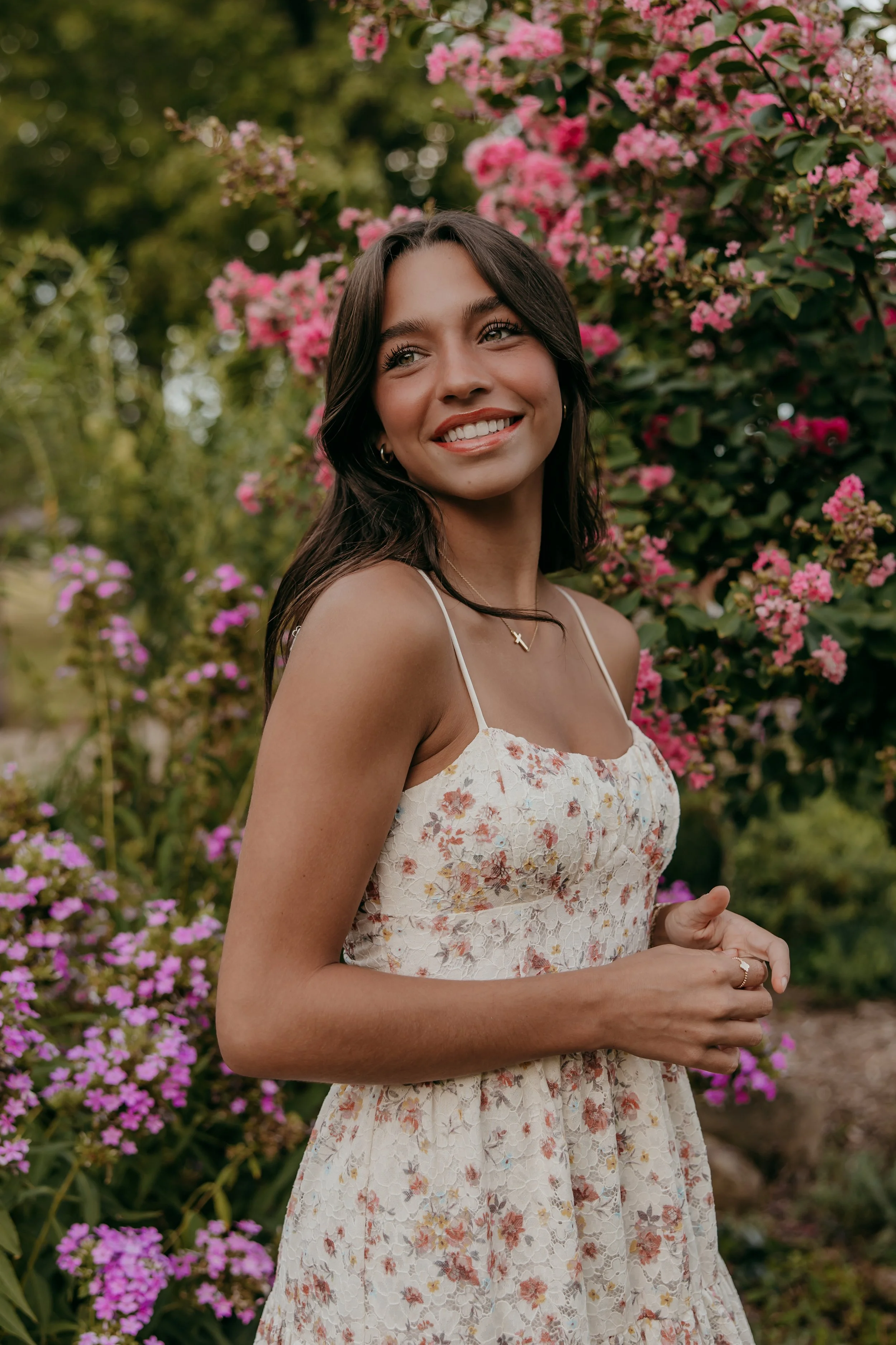 A young woman with long dark hair and light skin is smiling and standing outdoors in front of pink flowering bushes, wearing a white floral dress with spaghetti straps.