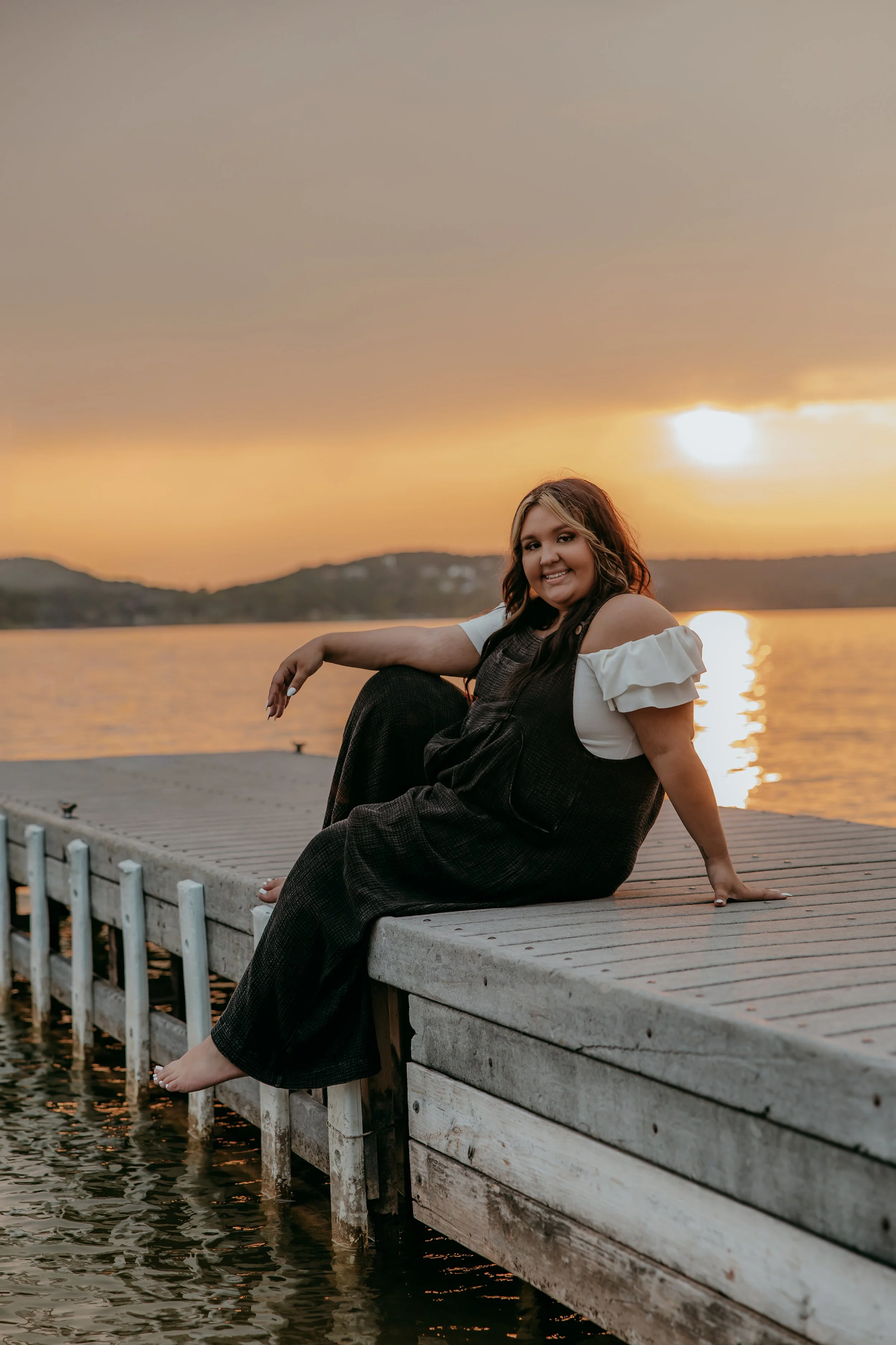 A woman sitting on a wooden dock by a lake during sunset, smiling, with hills in the background.