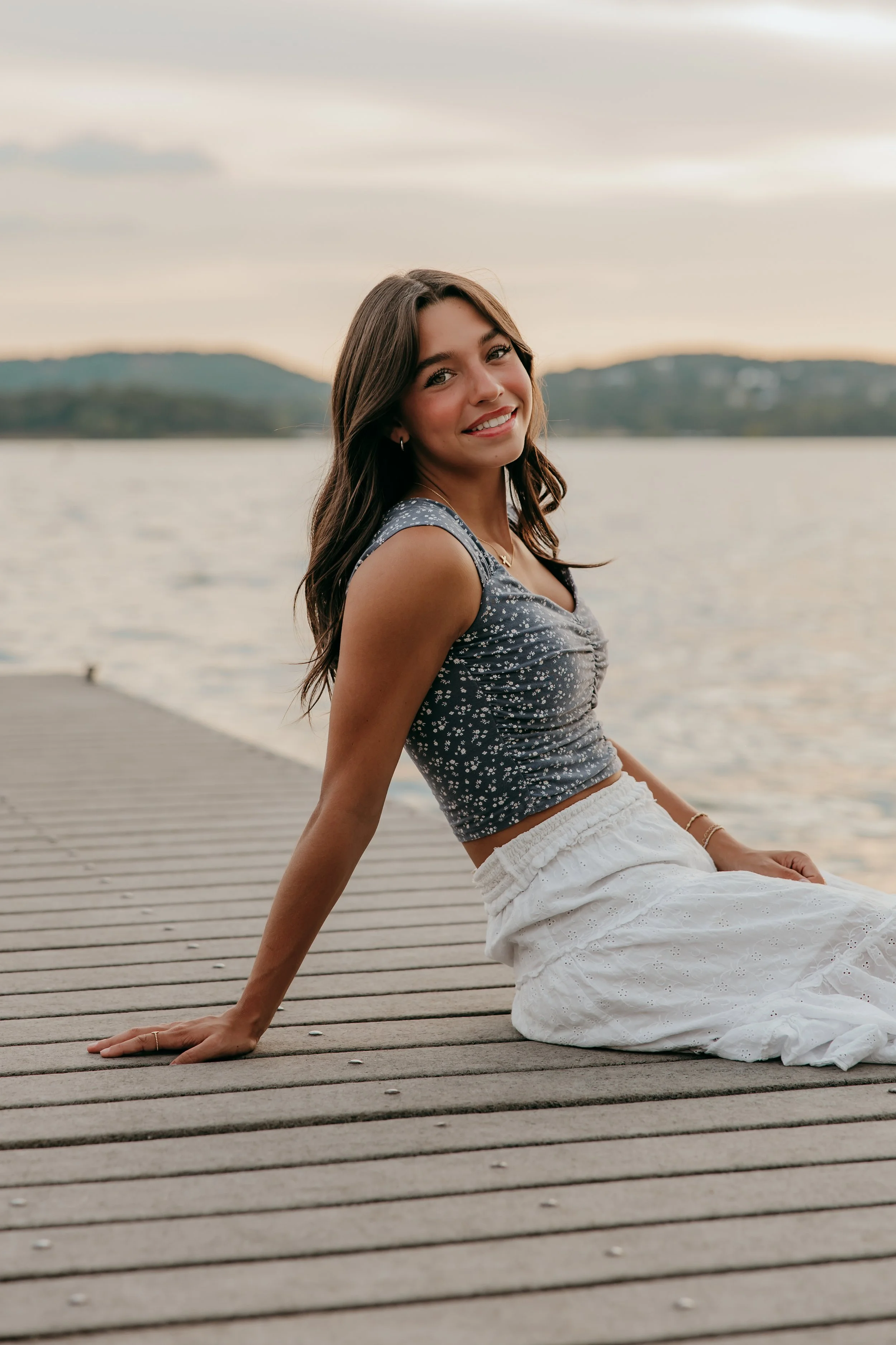 Young woman in a sleeveless floral top and white skirt sitting on a wooden dock by the water, smiling at the camera during sunset.