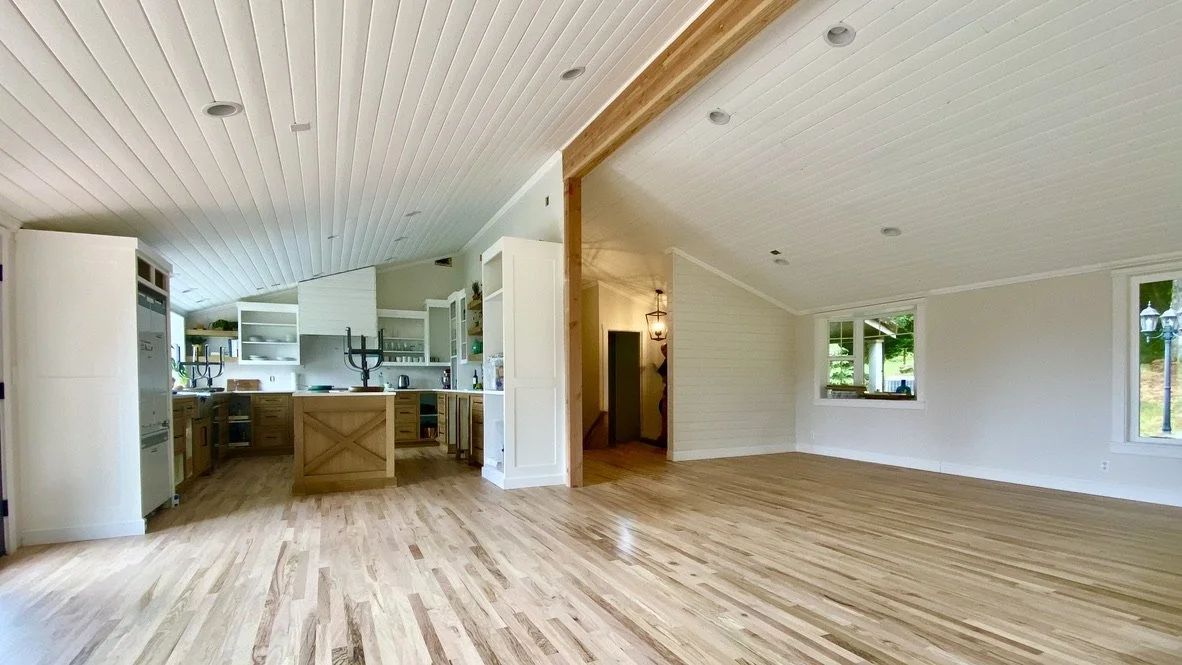 Open-concept living space with a kitchen on the left and empty room on the right, featuring white walls, wooden floors, and large windows.