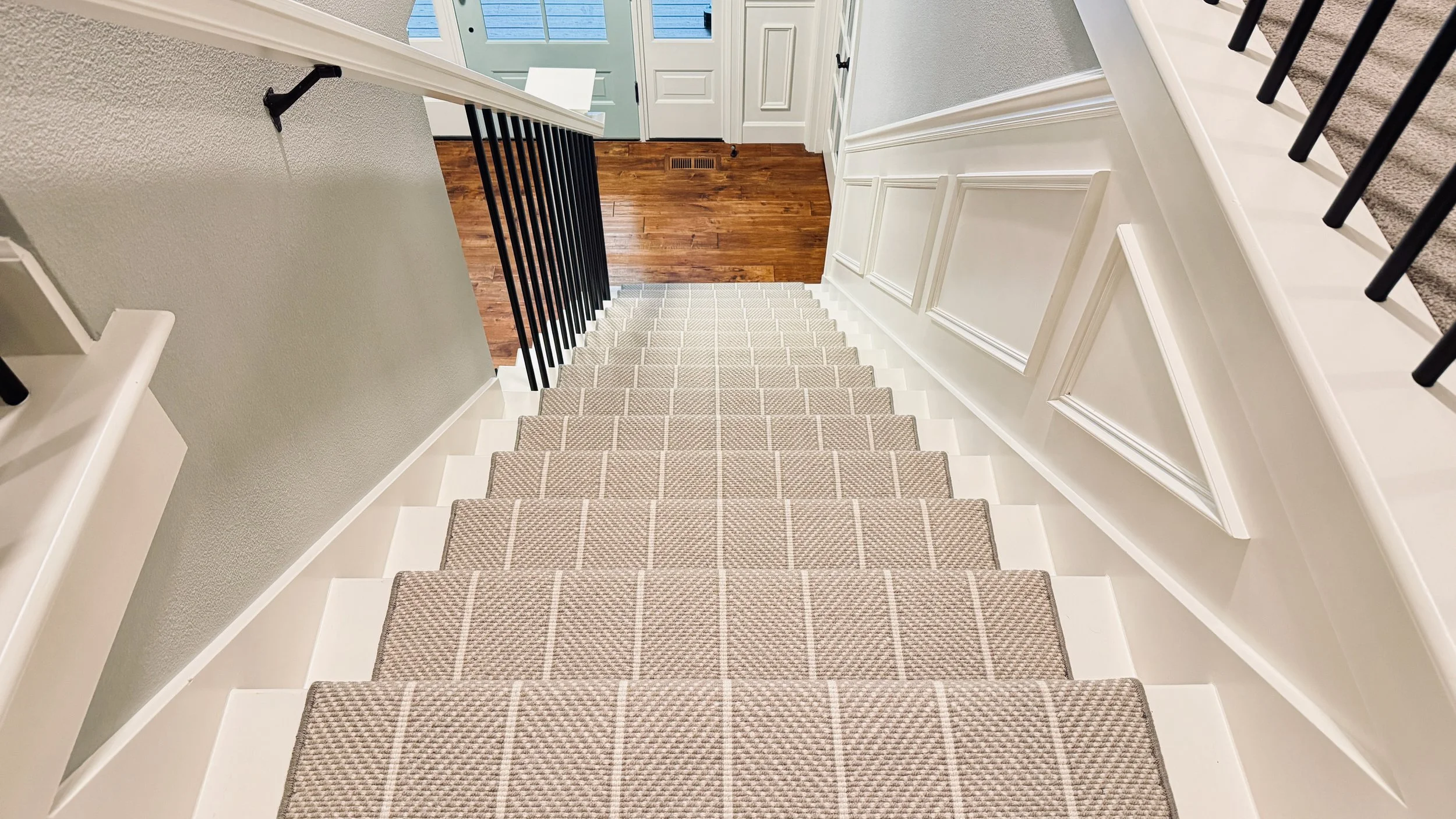 View of a staircase with beige striped carpet, white risers, black handrail on the left and white wood-paneled walls on the right, leading down to a hardwood floor entrance area with a front door and a window.