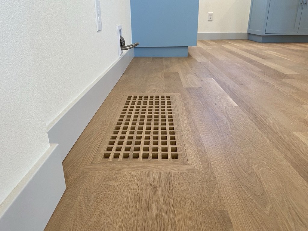 Wooden floor with a vent cover next to a white baseboard and a wall with outlets and a door.