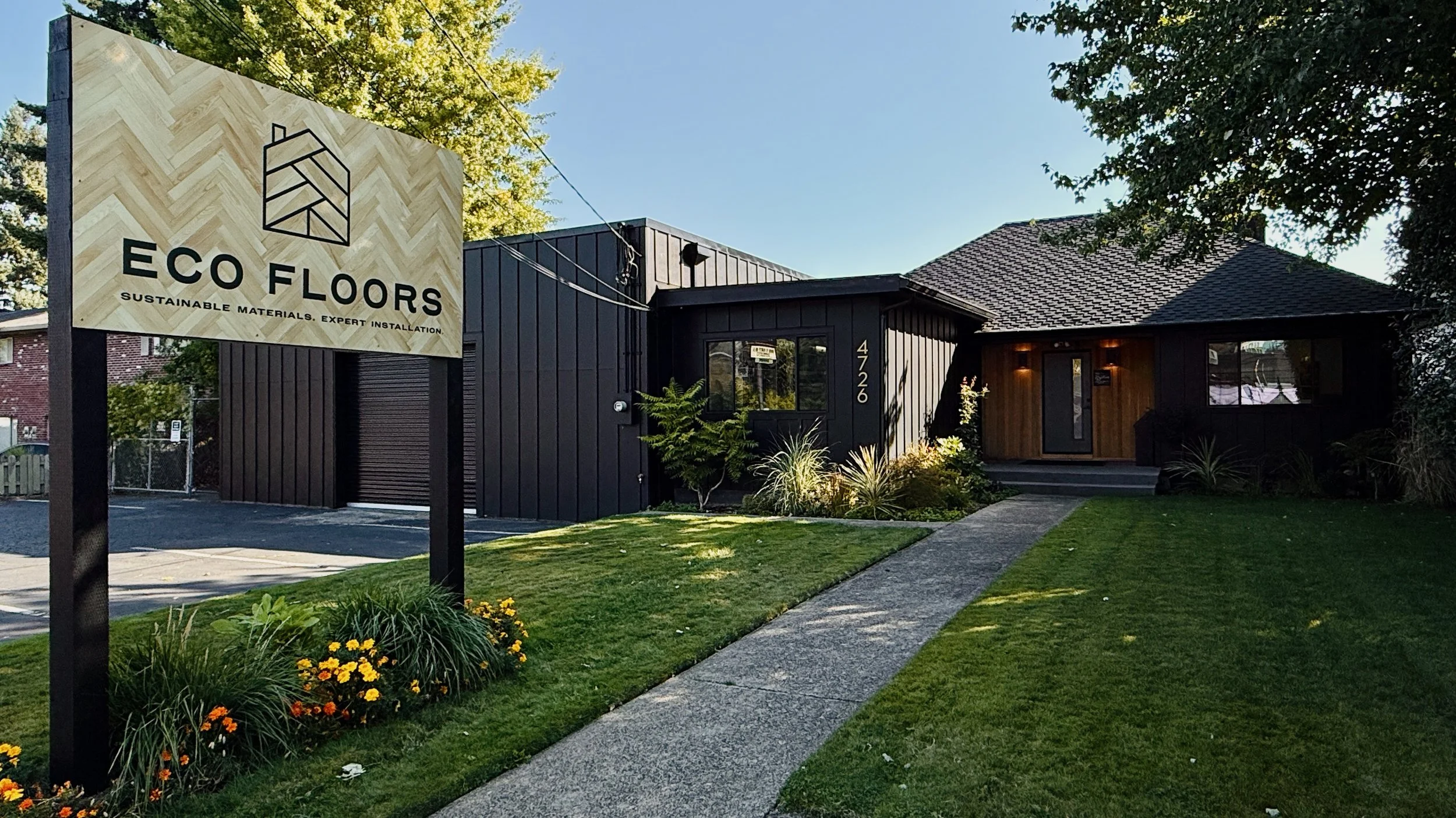 Eco Floors, Portland, OR Exterior - modern building with black and wooden siding, surrounded by a well-maintained lawn and flower beds, and a sign reading 'ECO FLOORS Sustainable materials, expert installation'.