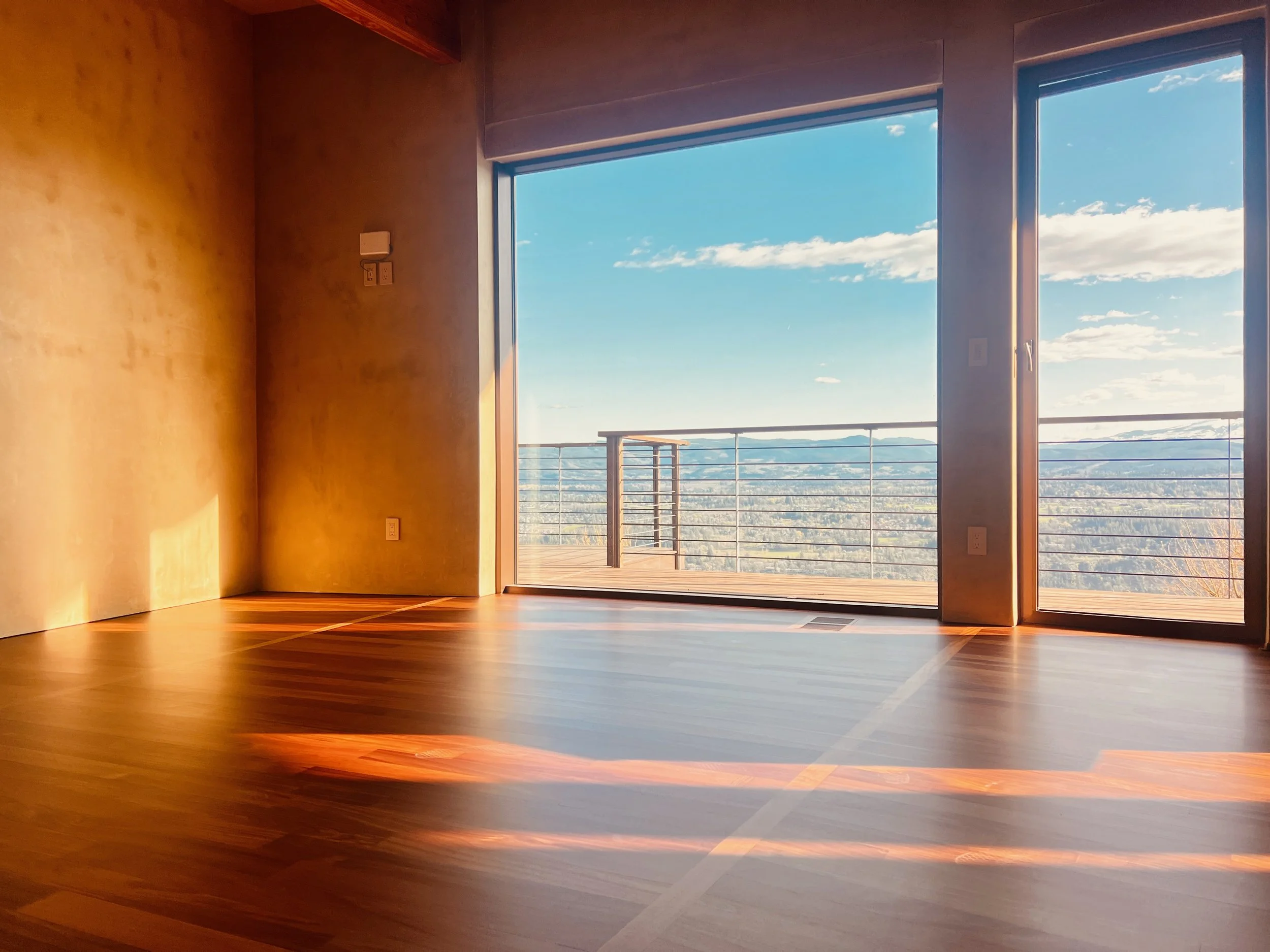 Empty room with cherry colored hardwood floors and large glass sliding window opening to a balcony with a view of Hood River, OR.