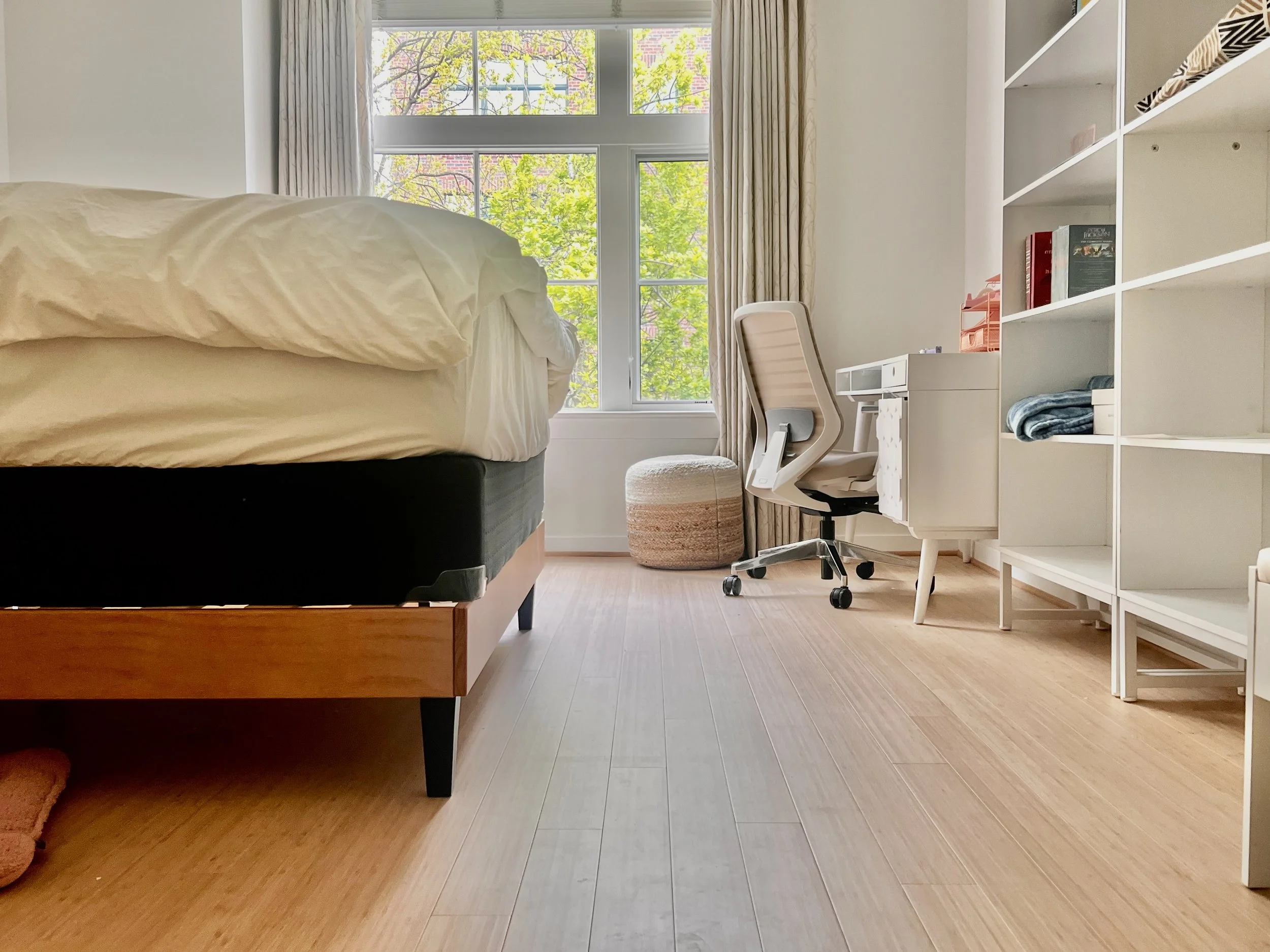 A bright, minimalistic bedroom with large windows showing green trees outside. The bed has white bedding, a white desk with shelves, a white office chair, and a round, woven ottoman on a light wooden floor.