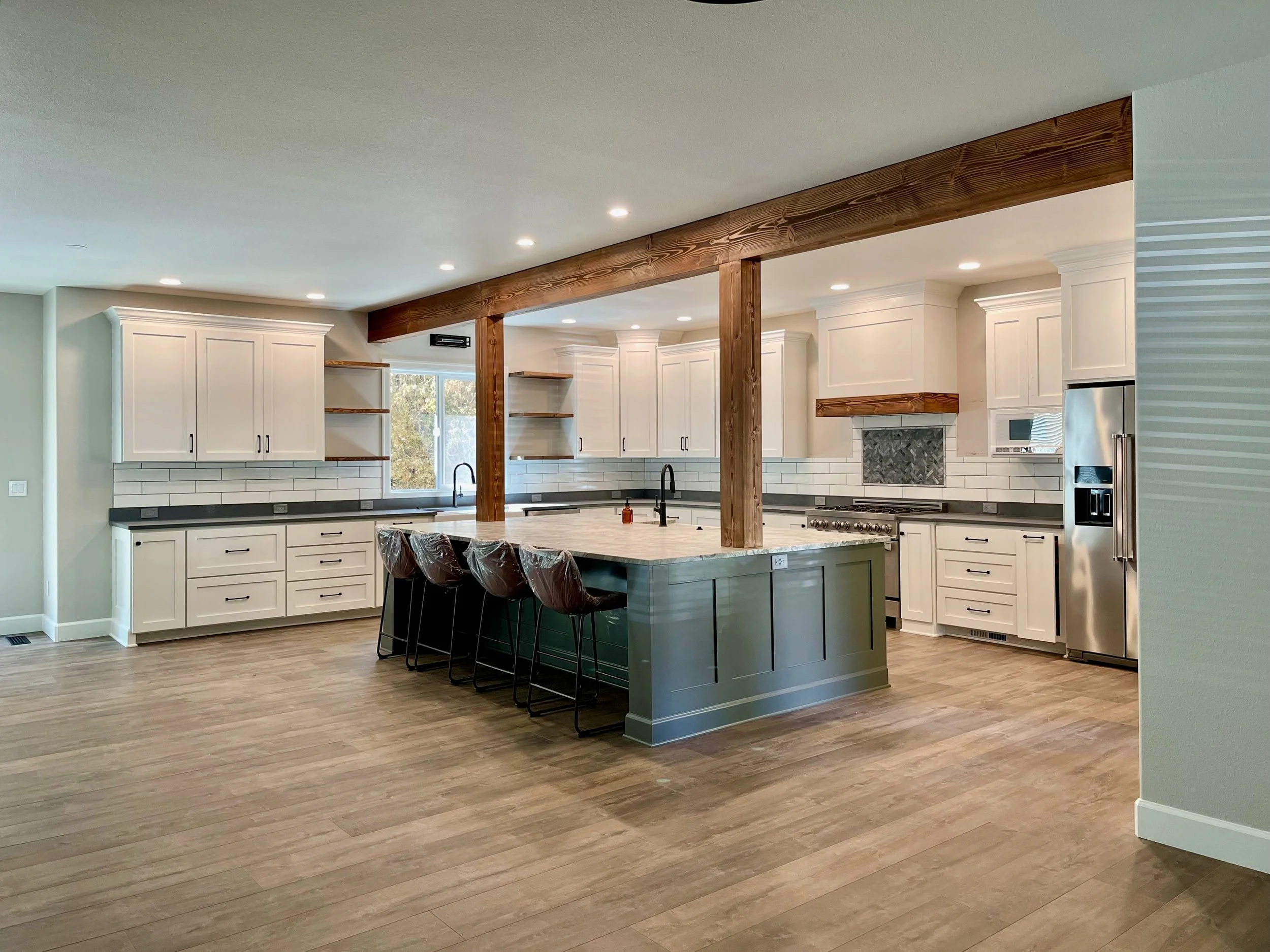 Modern kitchen with white cabinets, a large kitchen island with a marble countertop, wooden beams, stainless steel refrigerator, and a window letting in natural light.