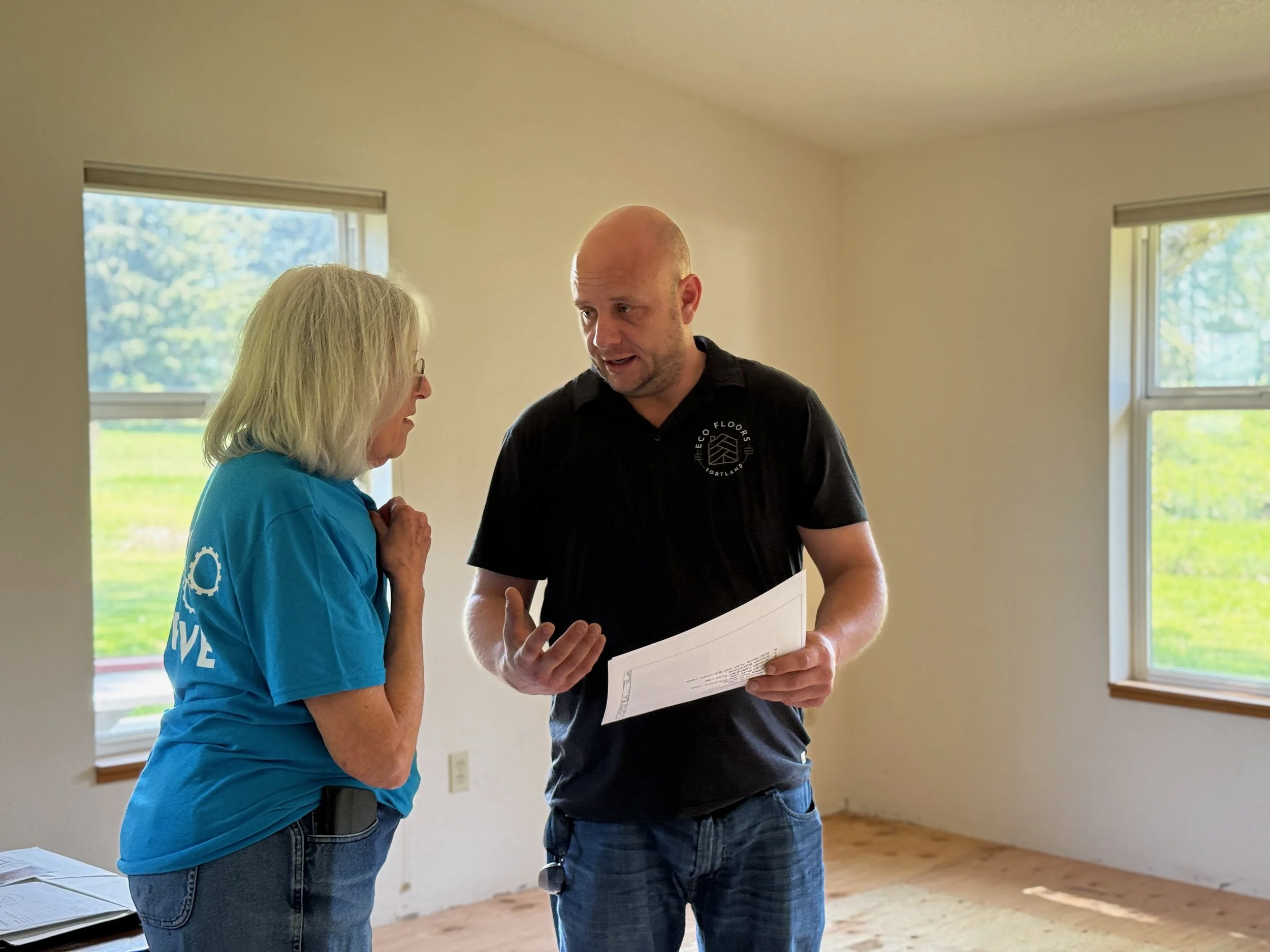 A man and a woman are discussing details of a home remodel inside a room with large windows and natural light. The woman is wearing a blue shirt, and the man is wearing a black shirt with an eco floors logo and holding a piece of paper.