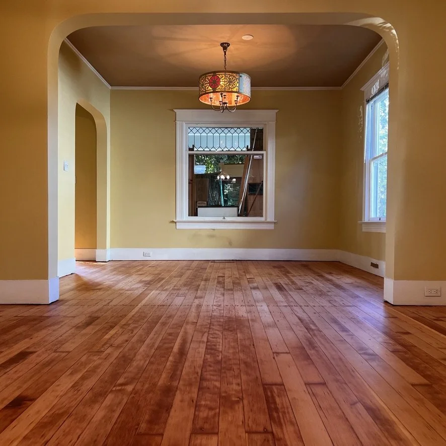 Empty living room with yellow walls, wooden floor, large window, and a colorful chandelier hanging from the ceiling.