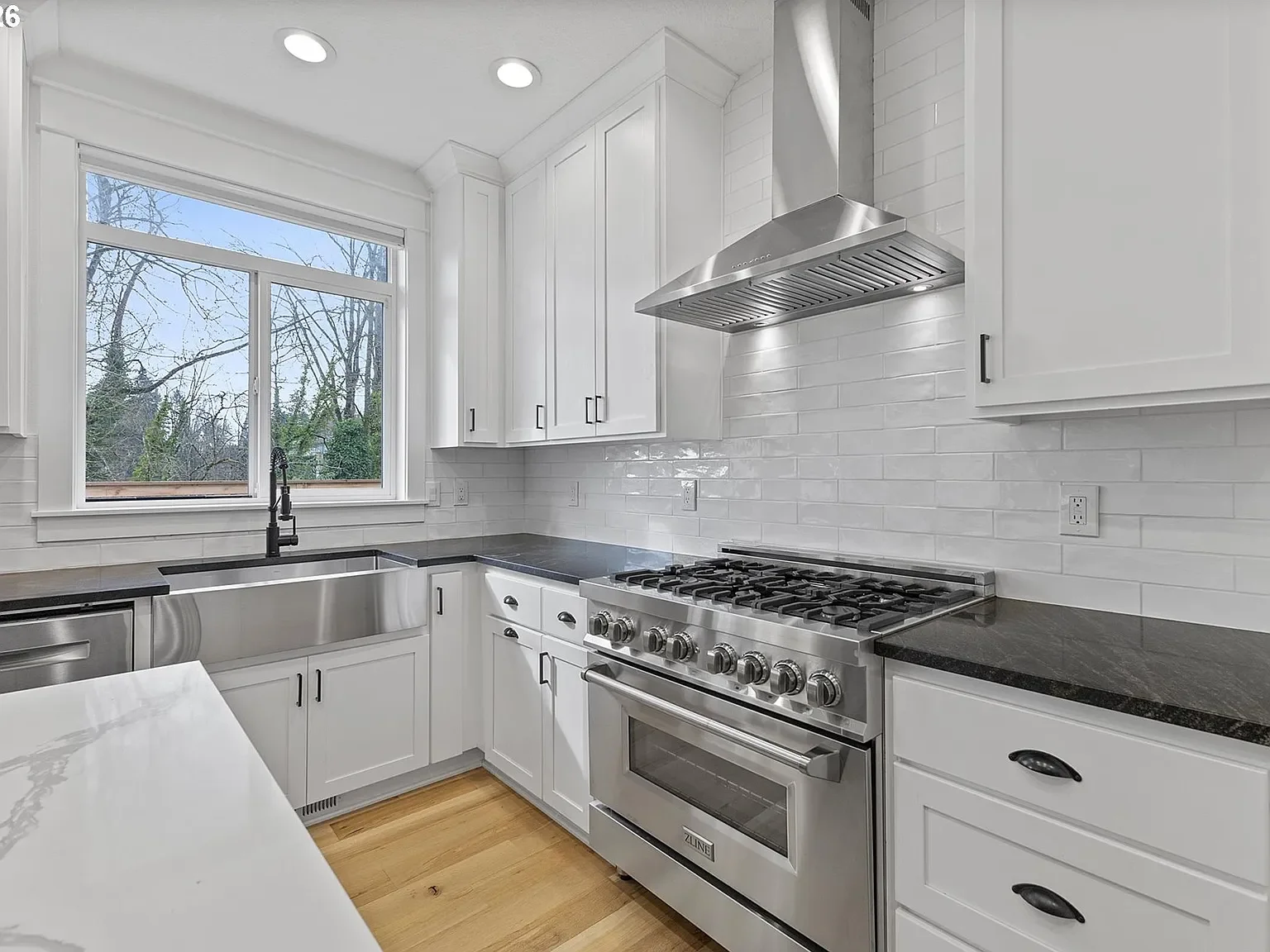 Modern kitchen with stainless steel range, hood, and sink, also featuring white cabinets with black quartz, and white oak hardwood flooring.