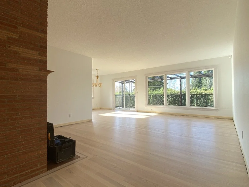 Empty living room with large windows, hardwood floors, and a brick fireplace on the left.