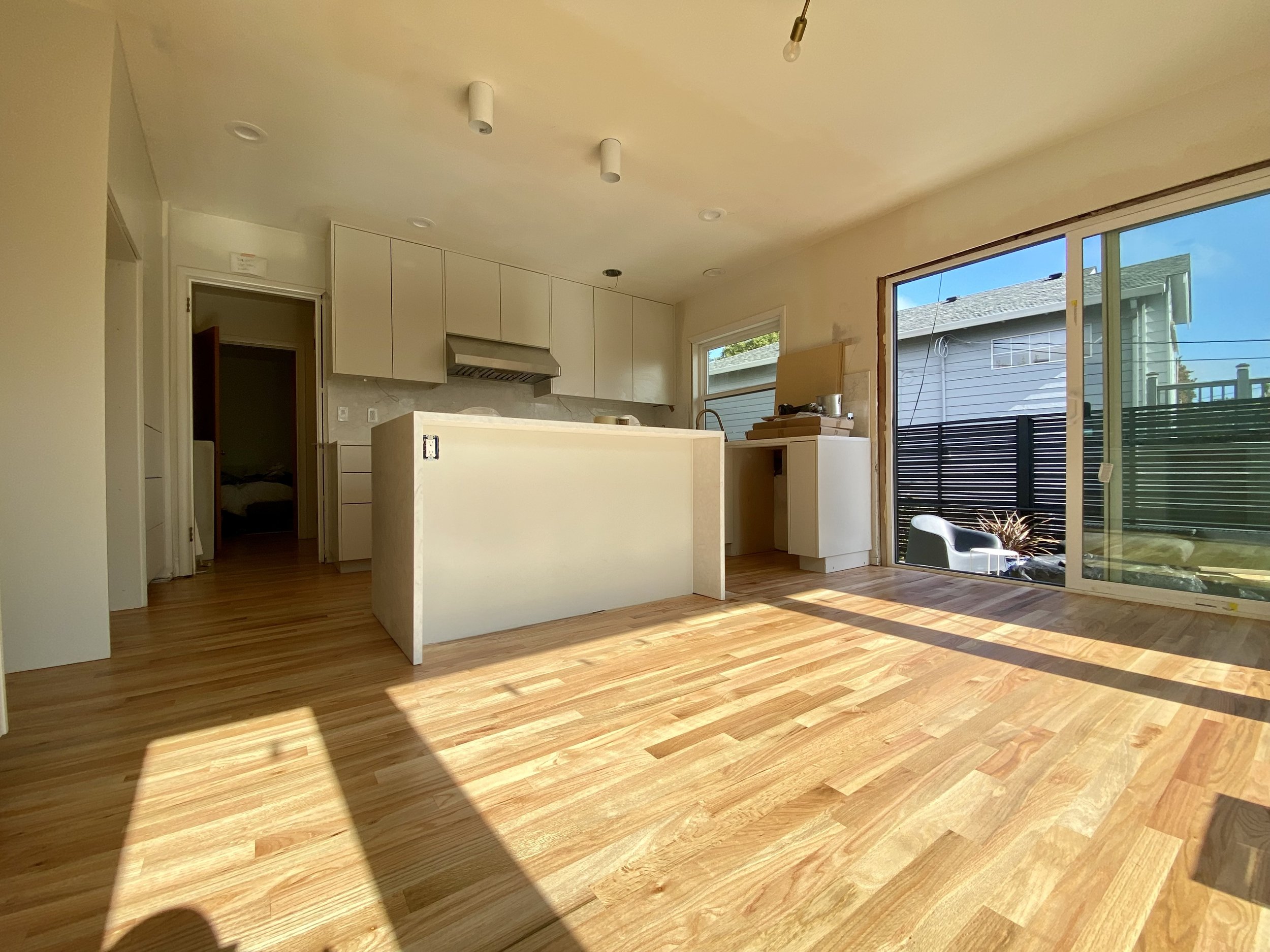 An interior view of a room with wooden flooring, white cabinets, a kitchen island, and large sliding glass doors leading outside. Natural sunlight illuminates the space.
