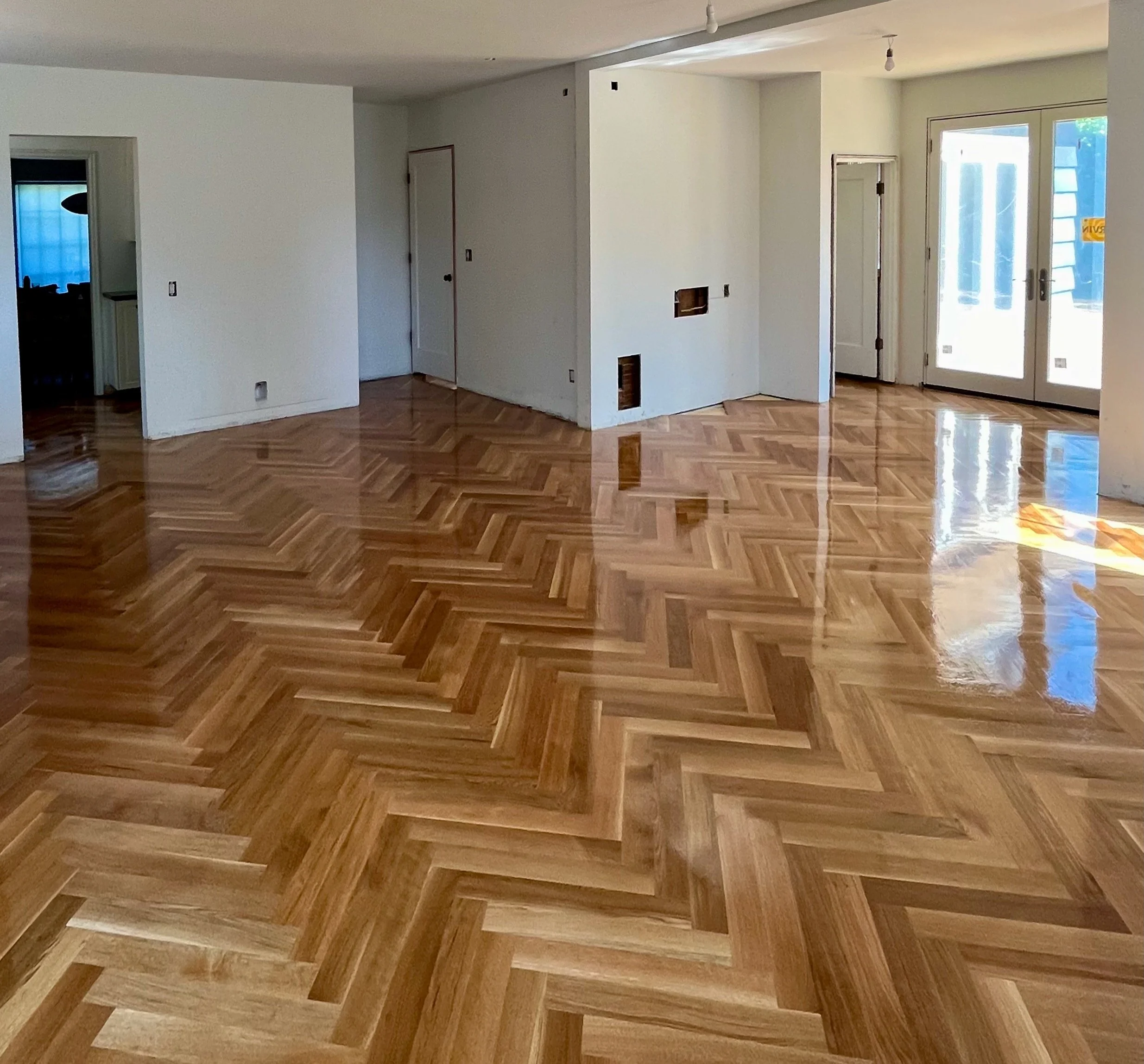 Interior of a house under construction with white walls, several doors, and a large window showing a blue sky outside. The floor has polished wood parquet flooring.
