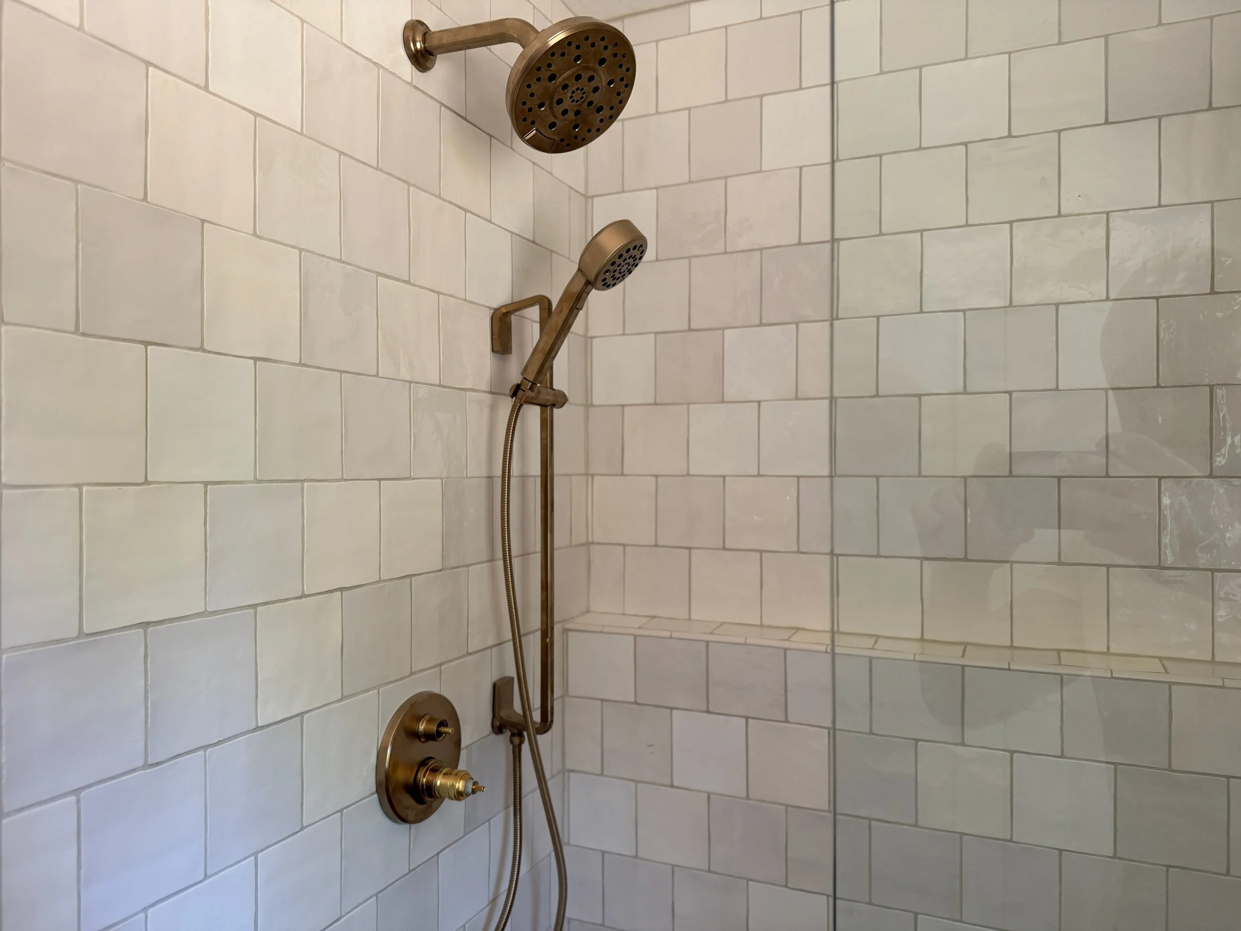 Shower with white natural edge subway tiles and bronze fixtures including a round overhead shower and a handheld shower head.