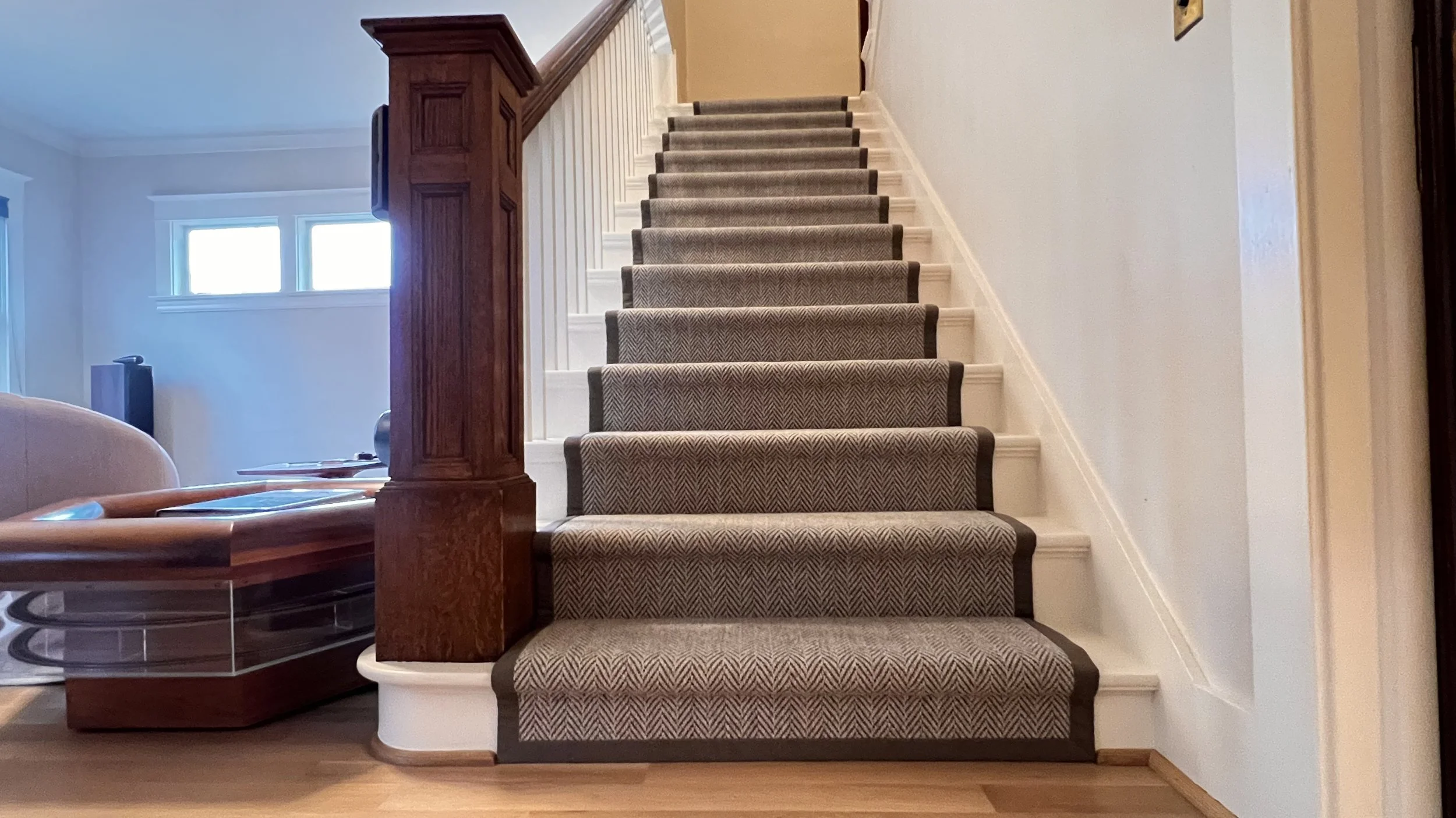 Interior view of a staircase with a chevron stair runner and wide edge binding, wooden handrail and built-in side table, next to a window in a house.