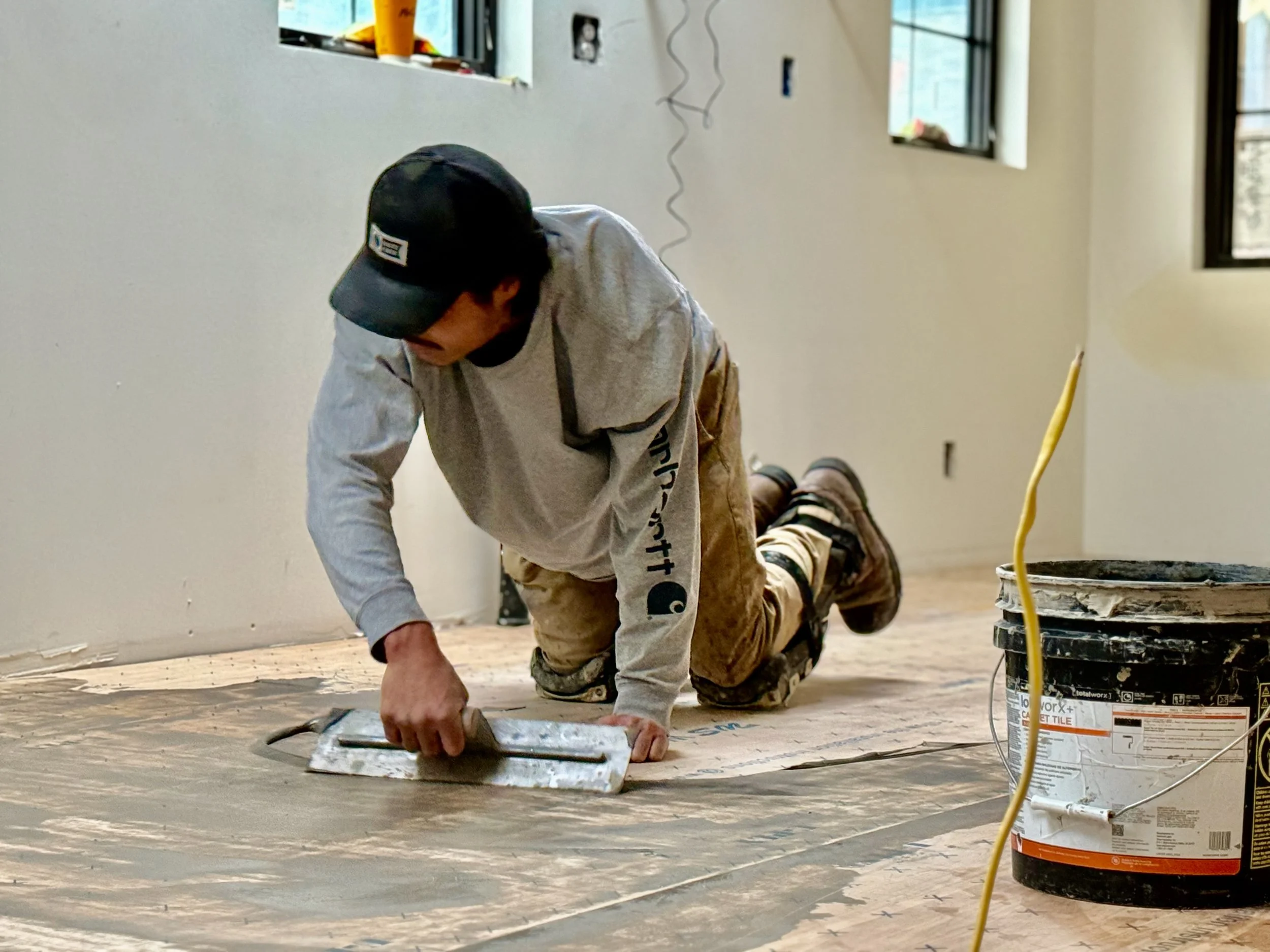 A person wearing a black cap and glasses is kneeling on the floor, applying a white substance with a trowel. They are working in a room with windows and unfinished walls, with a bucket of wall compound nearby, and construction tools and supplies around.