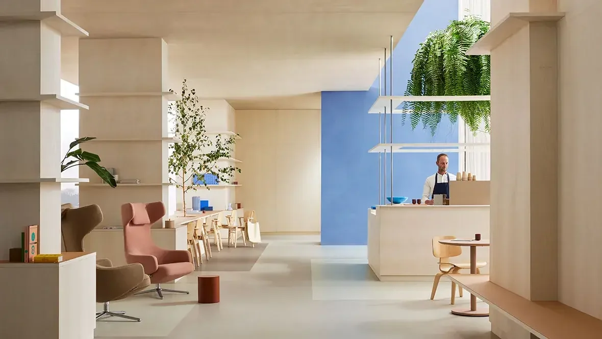 Minimalist cafe interior with light-colored walls, a counter where a barista is preparing drinks, and seating with modern chairs and tables. There are large green plants and natural light coming in.