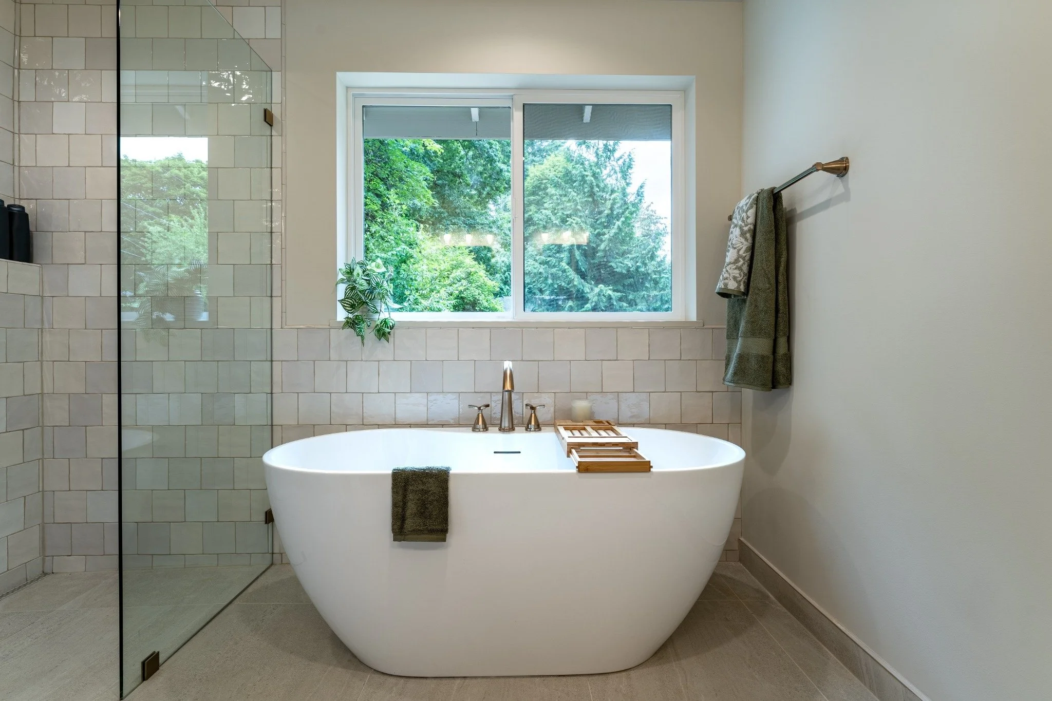 Modern bathroom with a freestanding bathtub, large window with a view of green trees, and a glass-enclosed shower on the left.