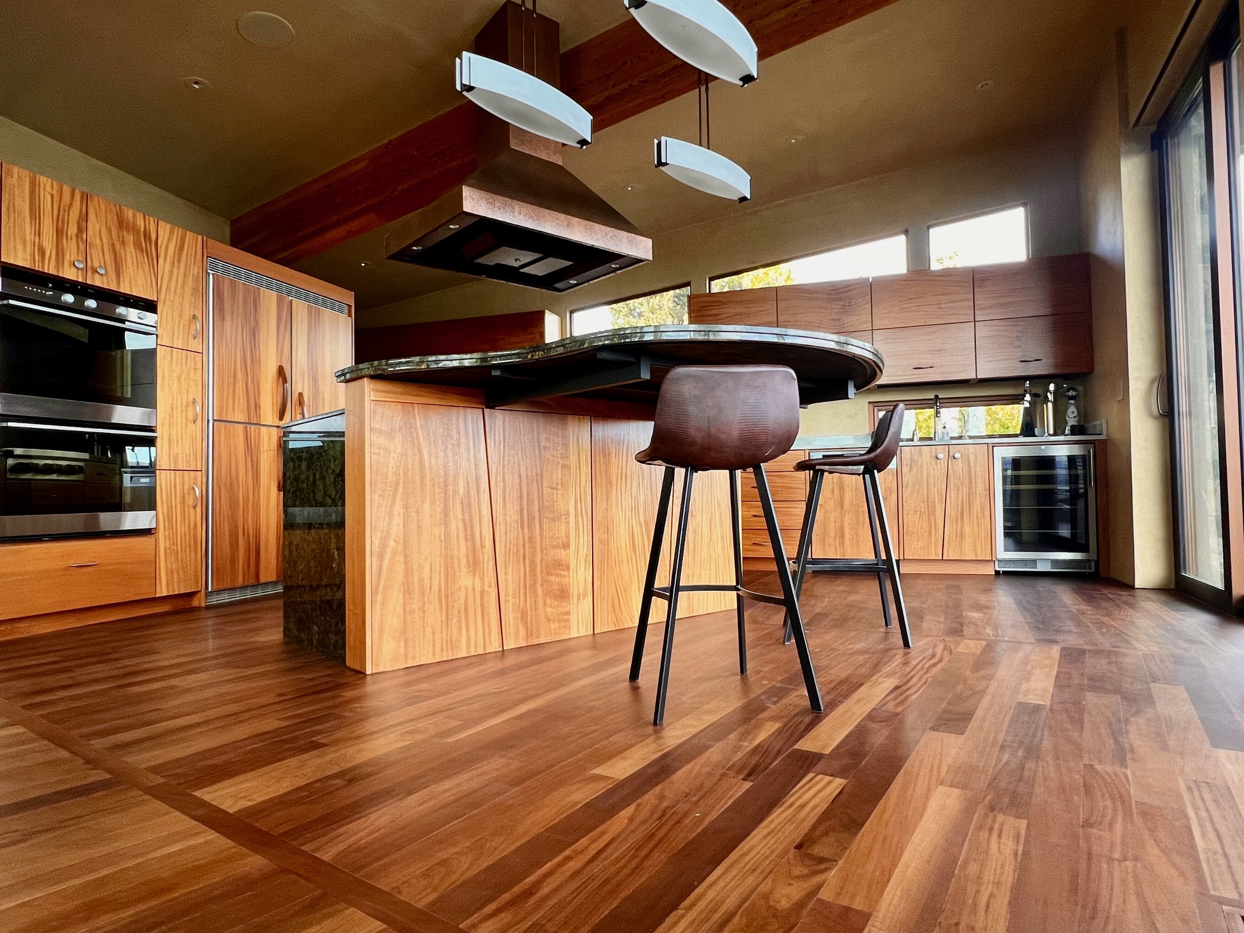 A modern kitchen with wood cabinets, a green marble-topped island, and high ceiling with hanging light fixtures. Large windows let in natural light.
