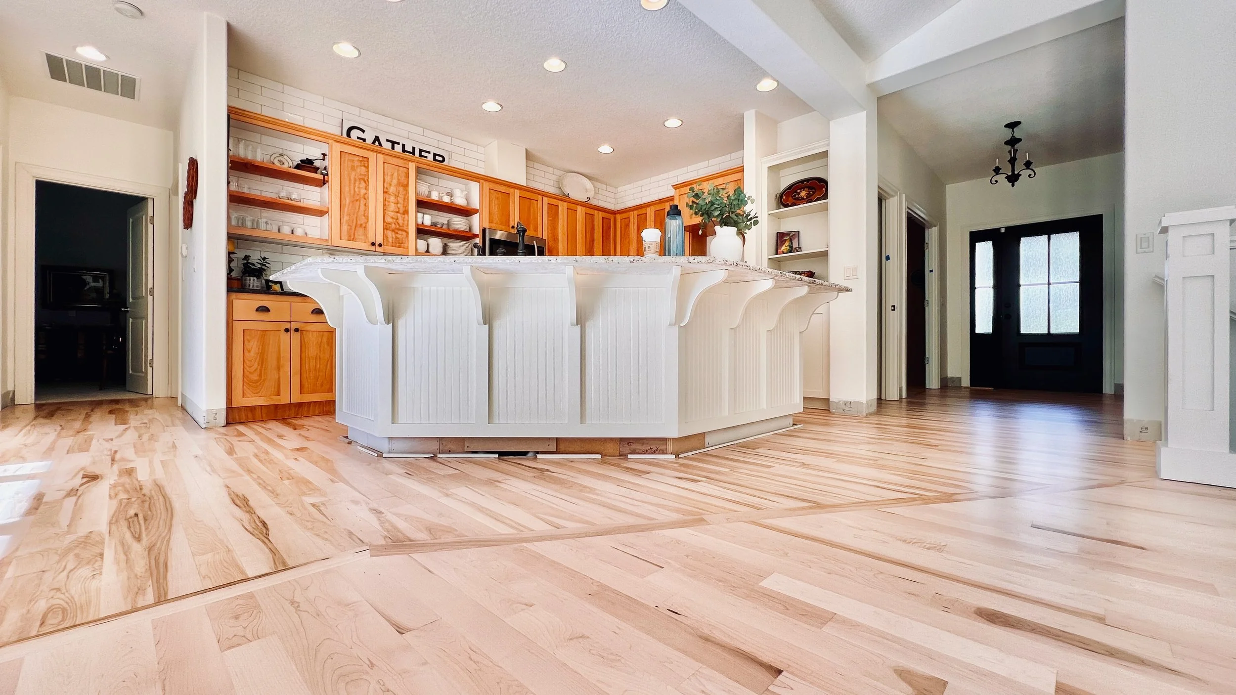 Open concept kitchen with light wood cabinetry and a large white island with decorative panels. Features include a built-in microwave, shelves with dishes, and a sign that says 'GATHER.' The space has light hardwood flooring, white walls, and recessed ceiling lights. There is an adjoining dining area visible to the right and a doorway to a room on the left.