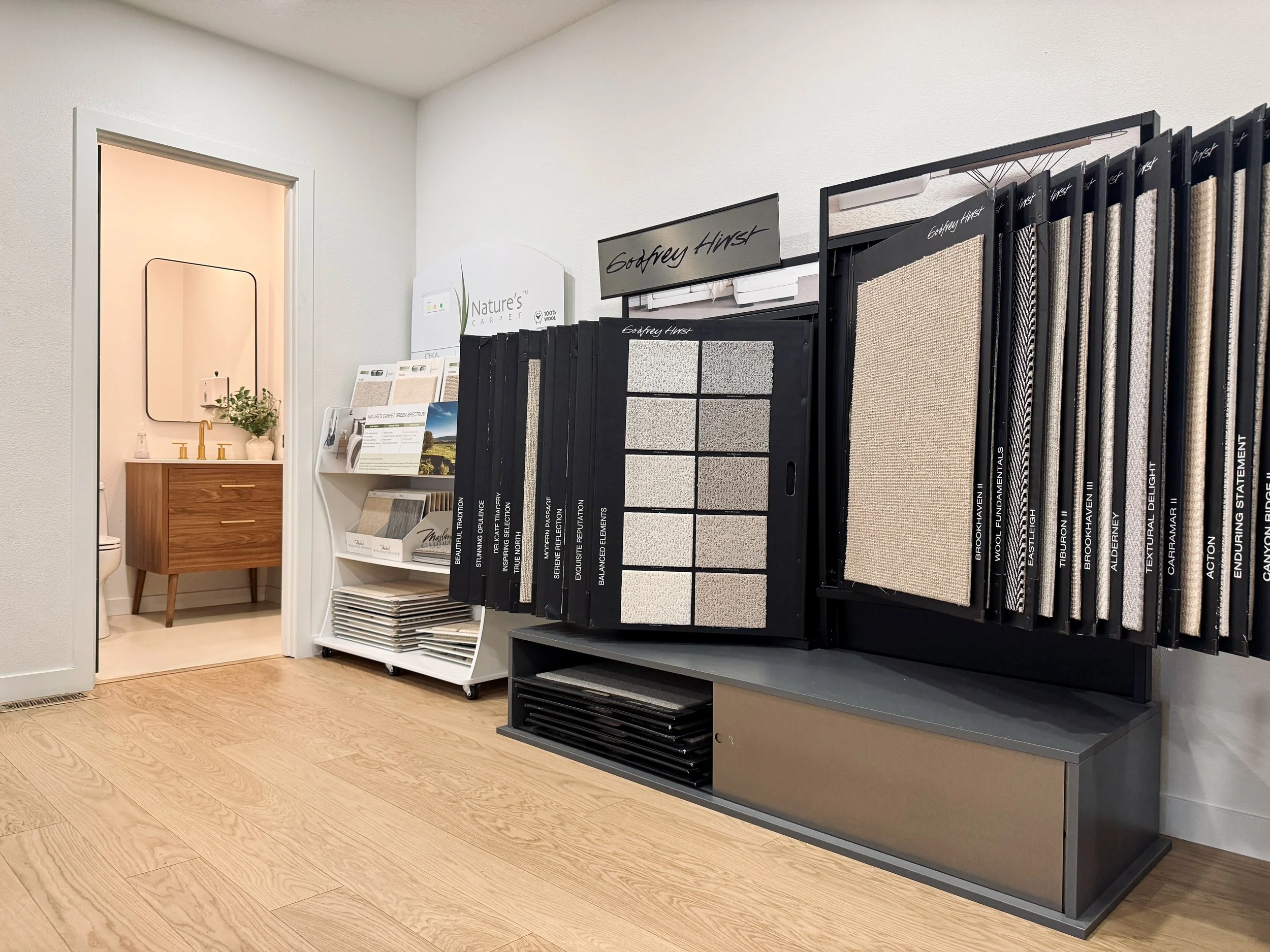Interior of a flooring store showing carpet and flooring samples on display racks, a small waiting area with a mirror, sink, and decor in the background.