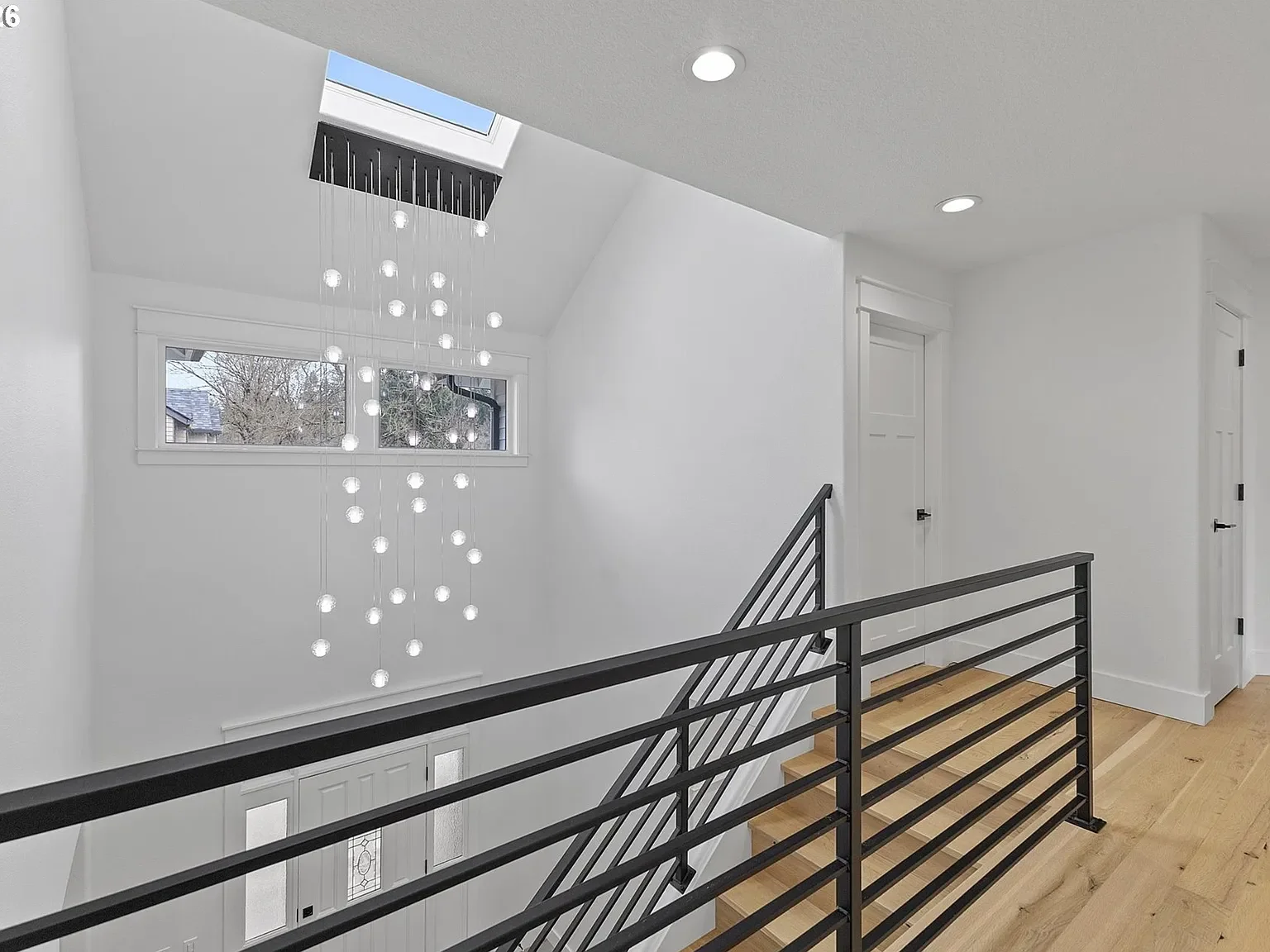 A modern hallway with a large glass globe chandelier, metal railing, and white oak flooring.