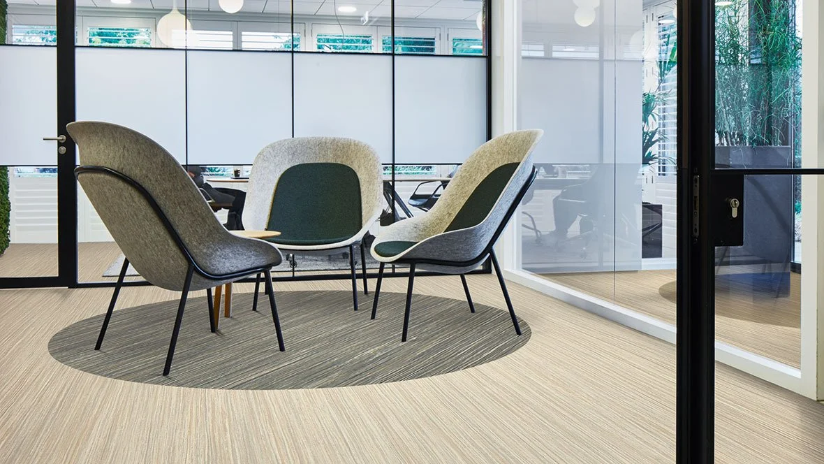 Modern office conference room with six cushioned chairs around a round table, glass walls, and a patterned rug, outside seating visible through the windows.
