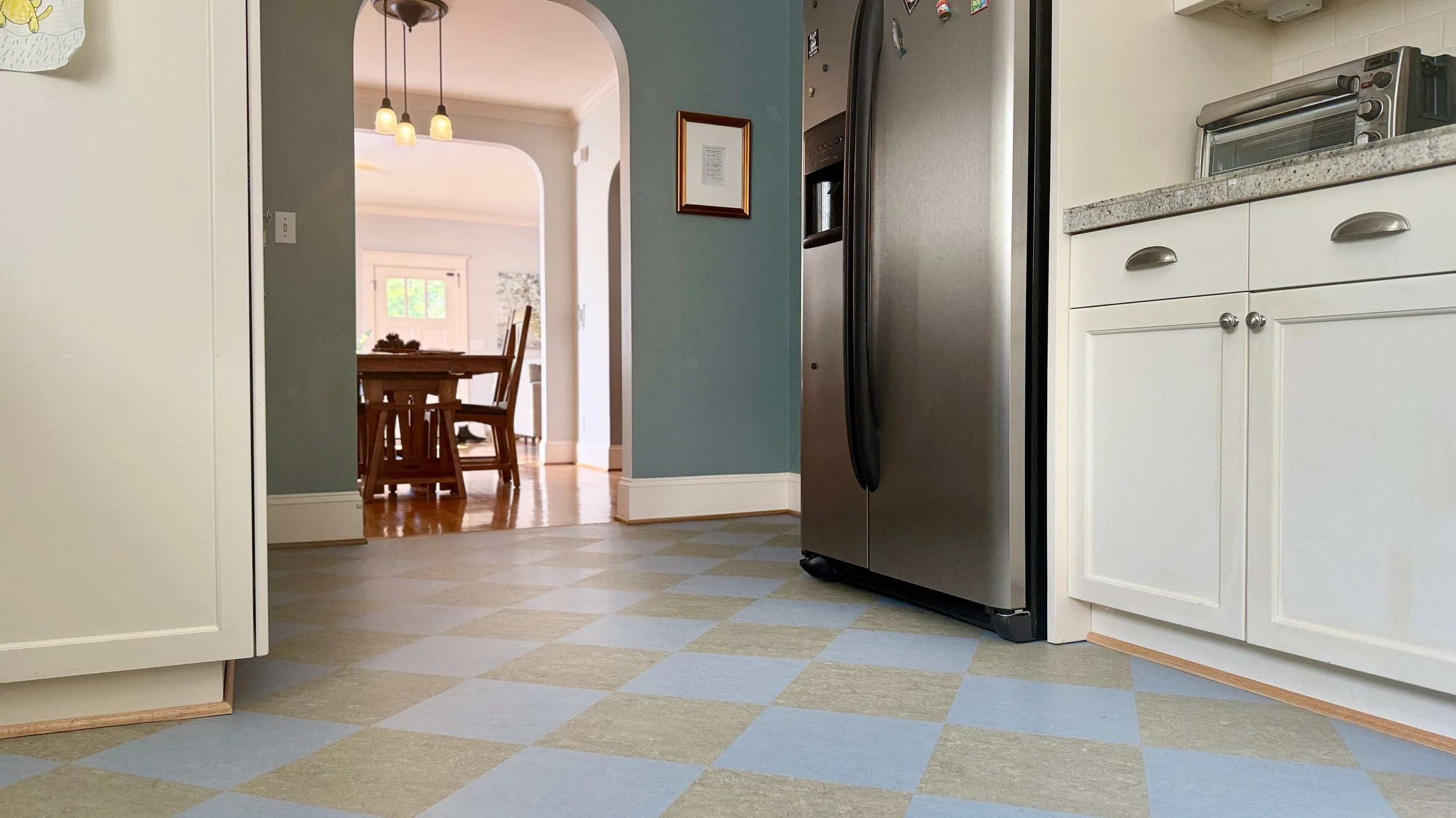 Kitchen floor with blue and beige checkered linoleum, white cabinets, stainless steel refrigerator, and a countertop toaster oven. A doorway leads to a dining area with a wooden table and chairs, visible in the background.