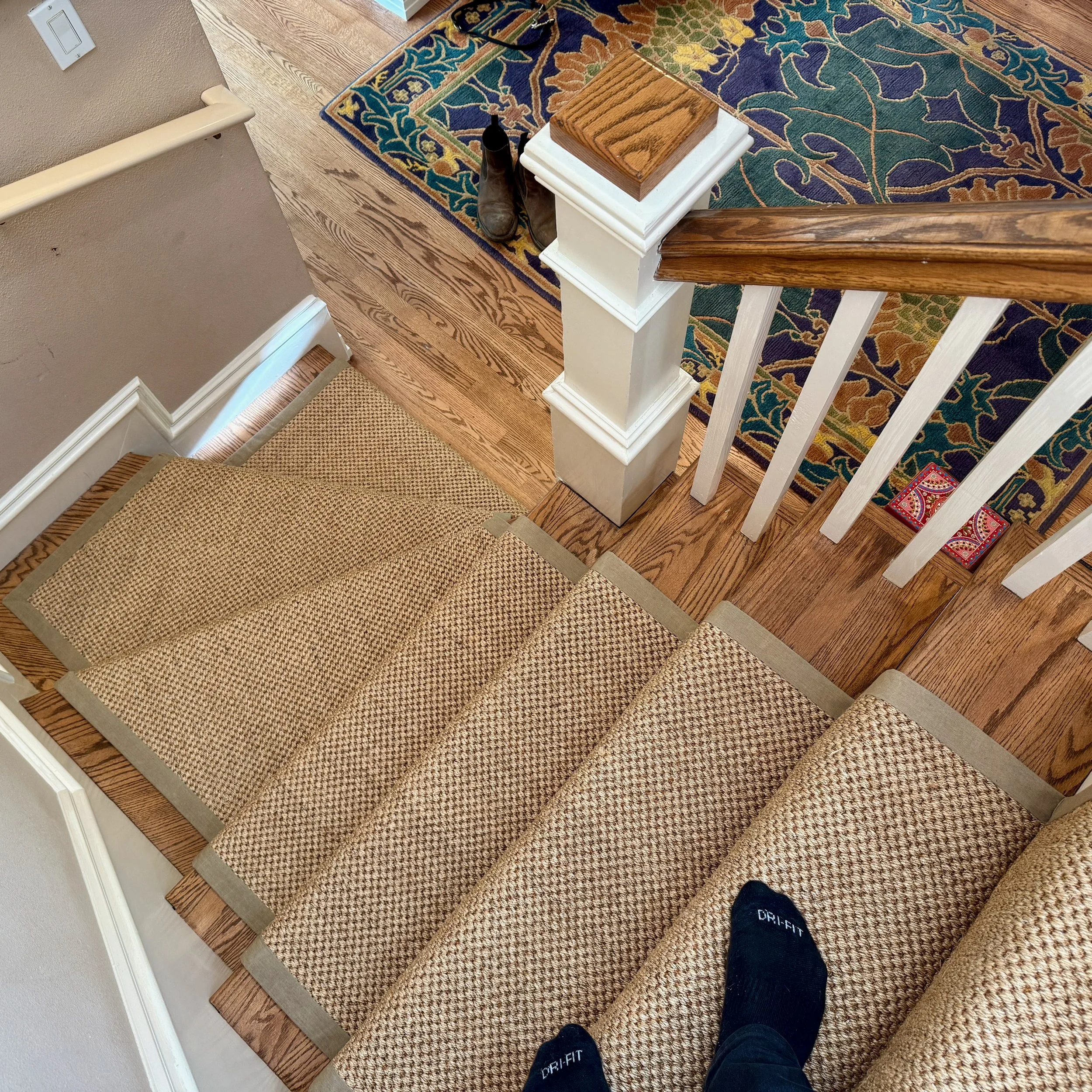 View of a wooden staircase with a carpet runner, showing a person's feet wearing black socks with white text, a wooden box on a white pedestal, a pair of black boots at the top of the stairs, and a colorful rug with floral design on a wooden floor.