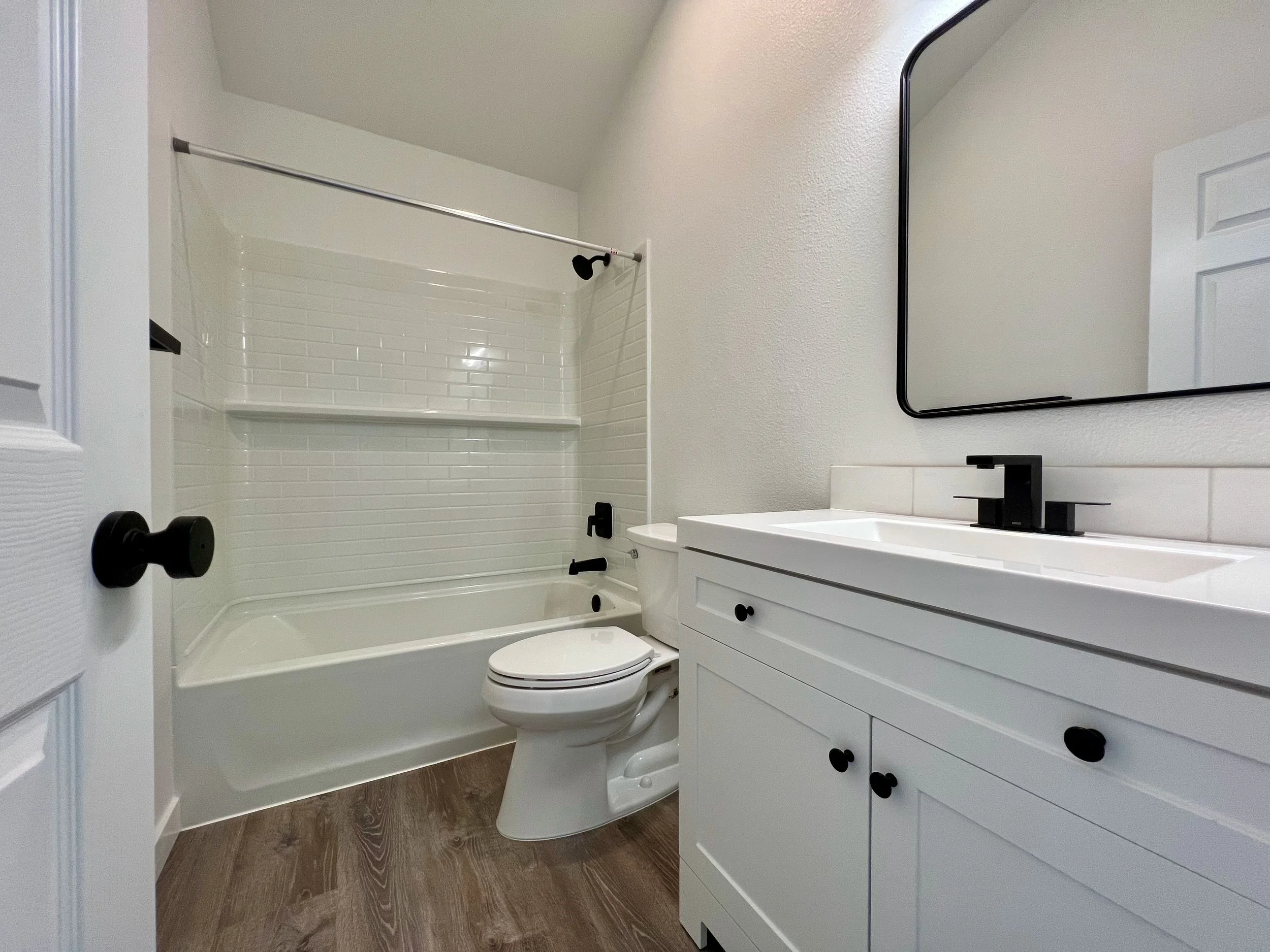 bright white restroom with laminate flooring, a subway tile style acrylic tub, and black fixtures. 