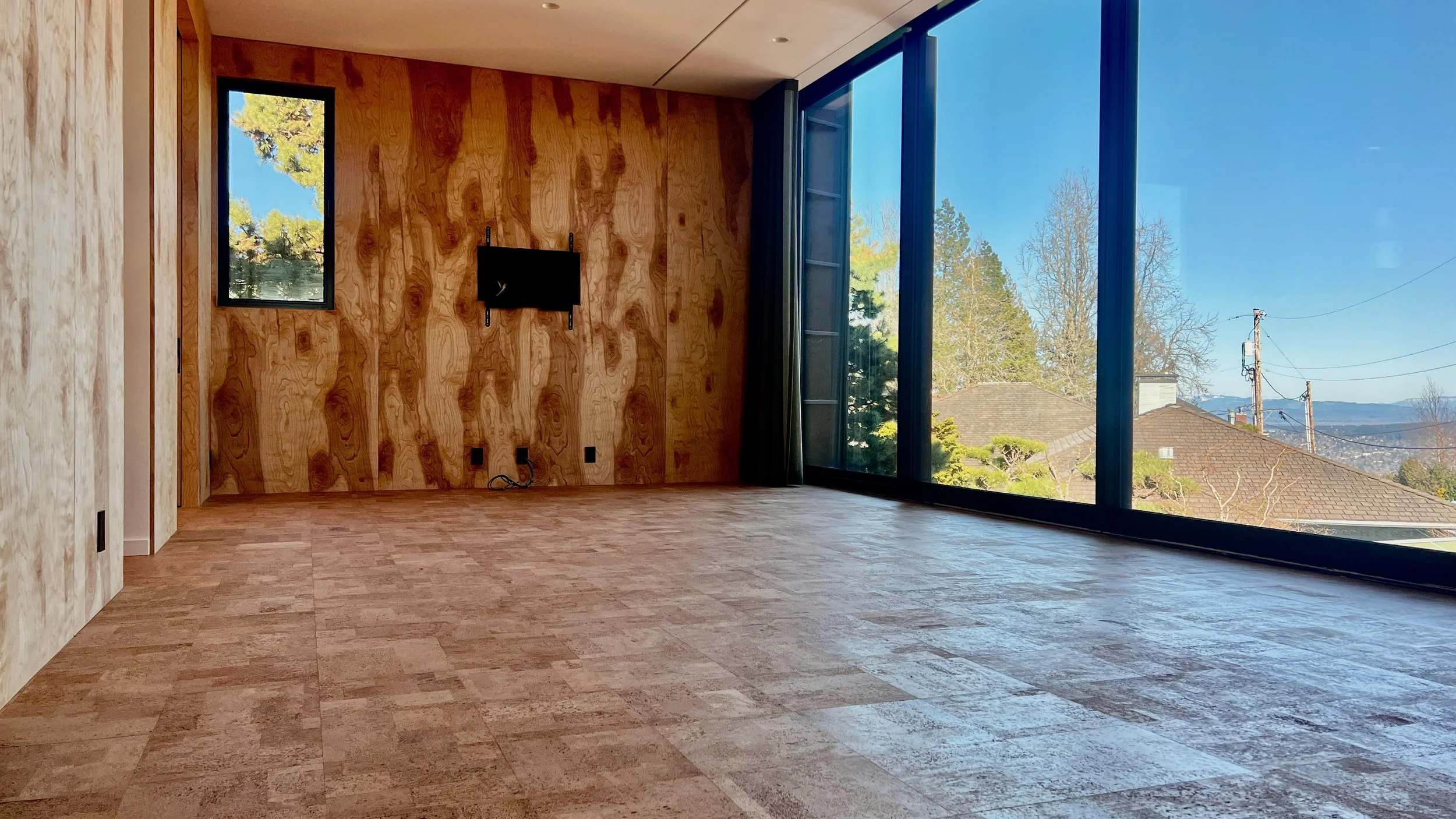 An empty room of a contemporary house with glued down, 1'x1' square cork tiles and a large window showing a scenic view of trees and rooftops.