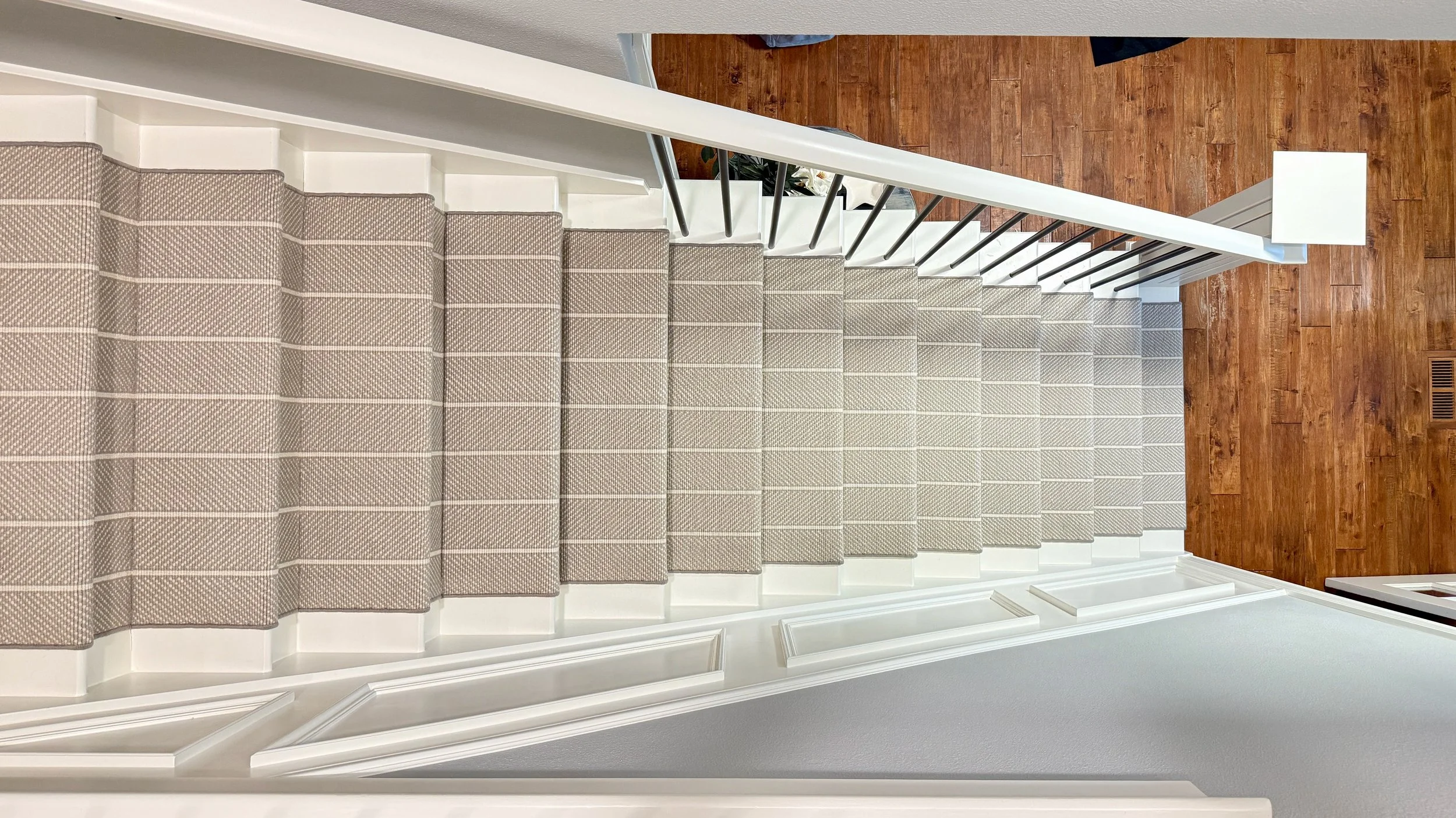 Top view of carpeted staircase with beige and white striped carpet, white stair risers, white banister with black spindles, hardwood floor at the bottom, and a portion of a white wall.