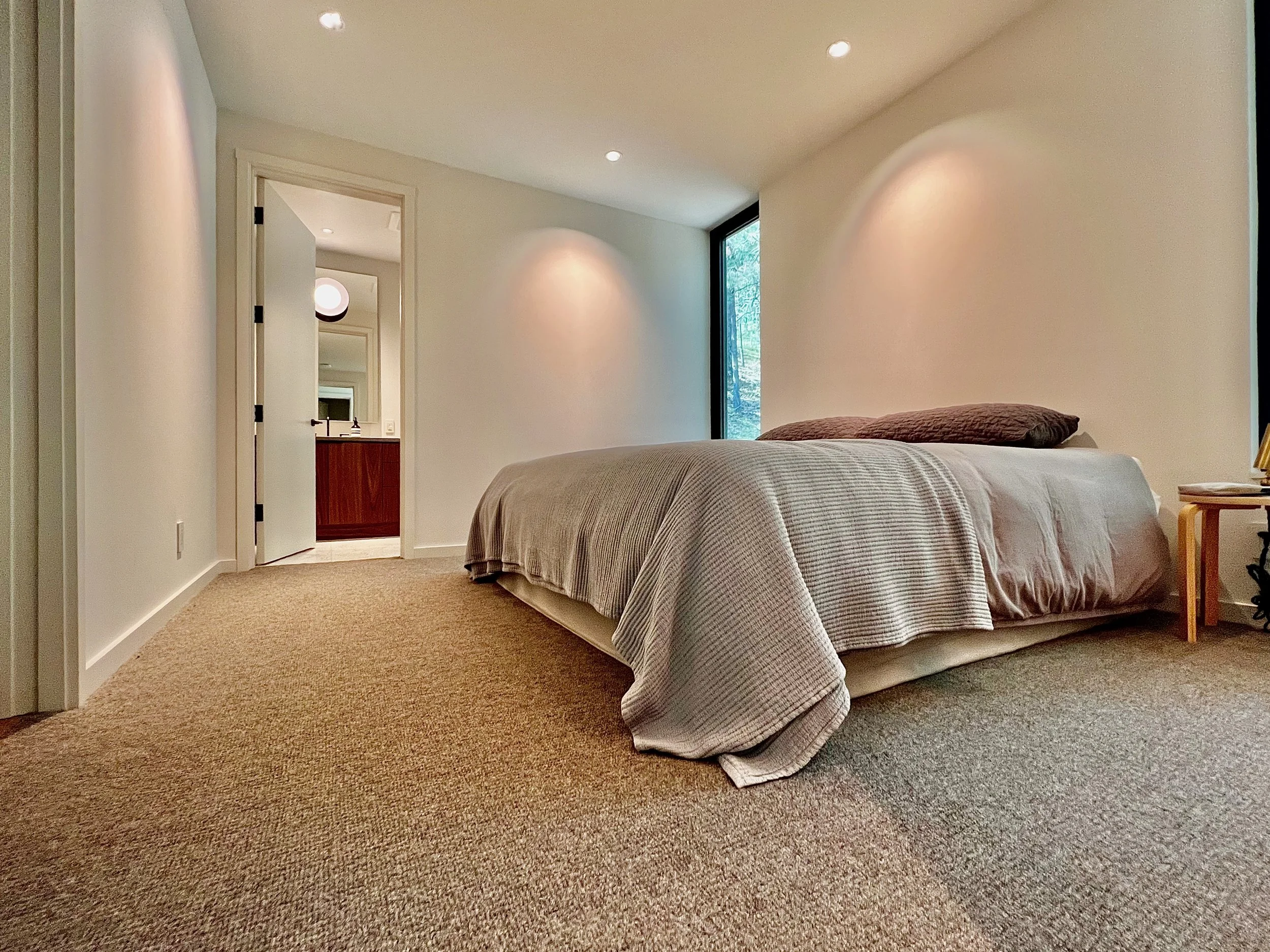 A bedroom with a bed covered in beige and white striped bedding, a brown pillow, beige carpet, white walls, a large window to the right, and an open door in the background leading to a bathroom with a round mirror.