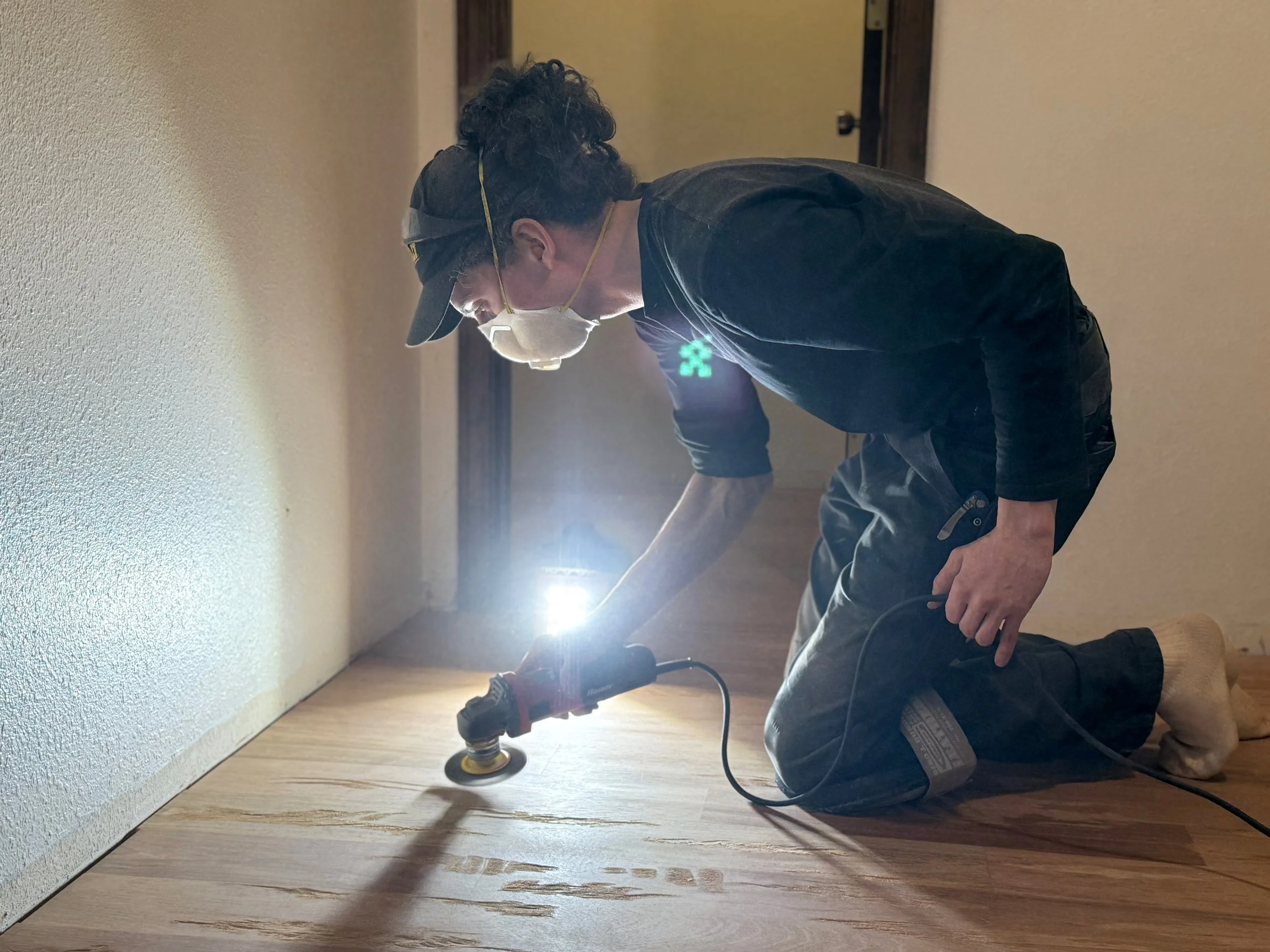 Man kneeling on the floor using a power sander on a hardwood floor near a wall, wearing a face mask and a cap.